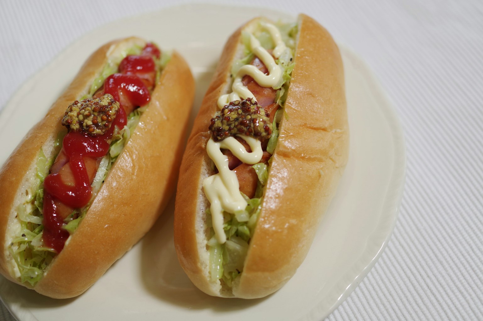 Dua hot dog di atas piring satu dengan saus tomat dan selada yang lain dengan mayones dan tomat