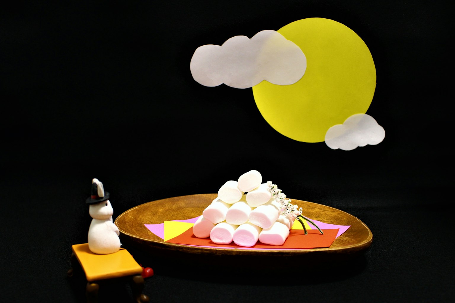 Craft scene featuring a yellow sun and white clouds on a black background with a wooden plate stacked with marshmallows