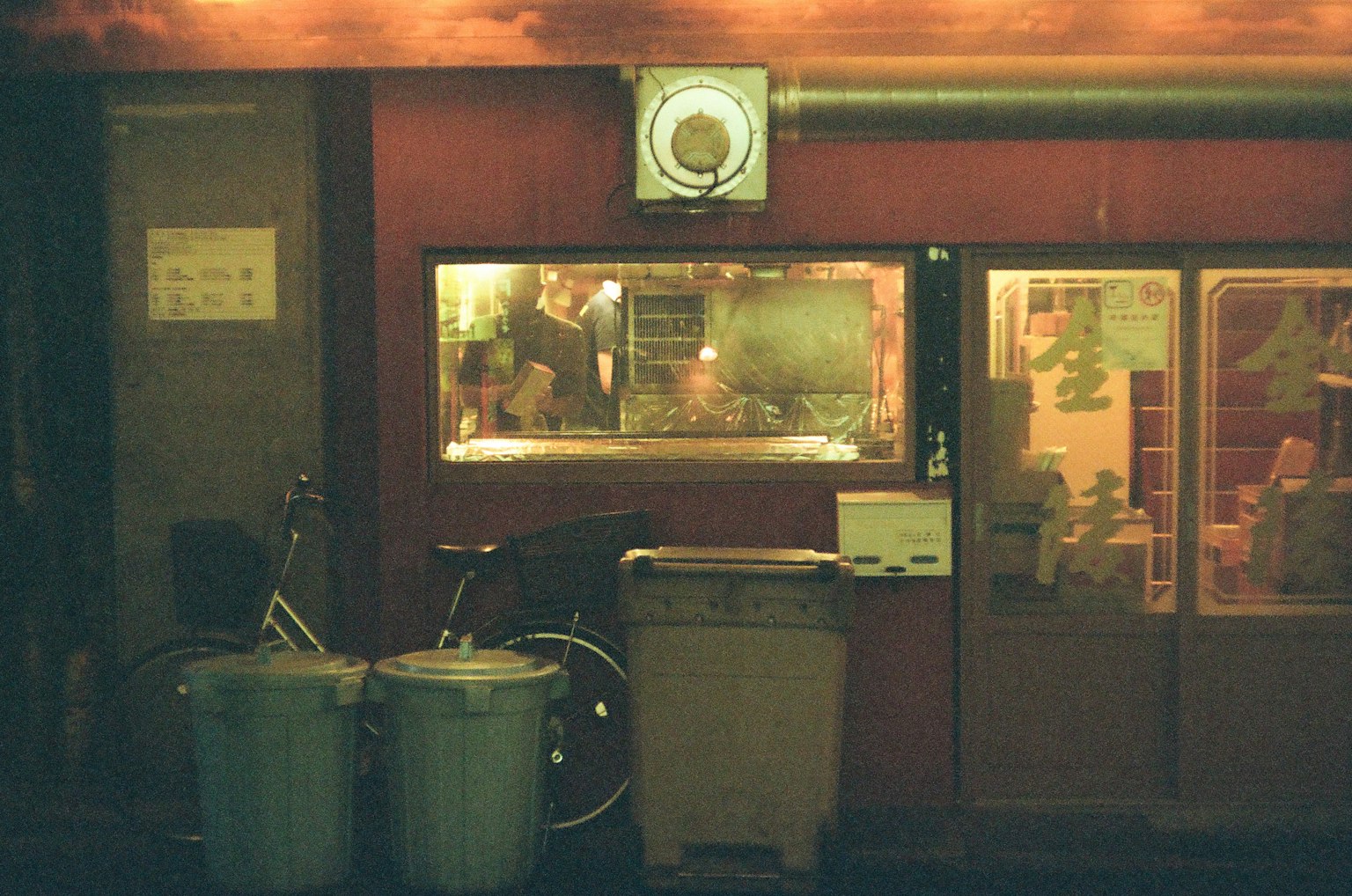Exterior of a small eatery at night featuring bicycles and trash bins