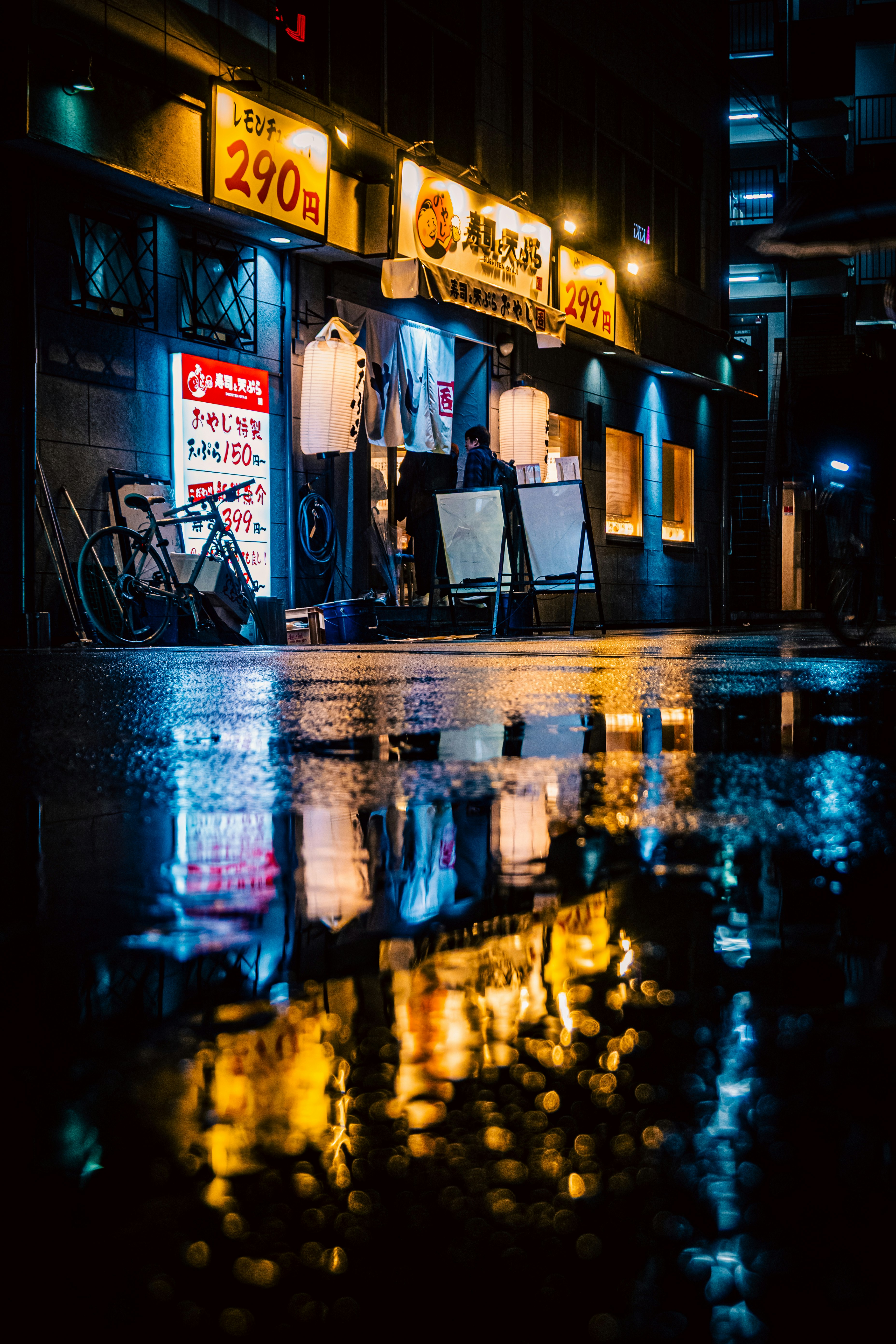 雨上がりの路地に並ぶ看板と自転車のある夜景
