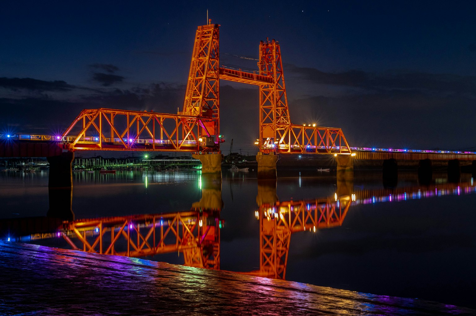 Jembatan yang diterangi dengan lampu berwarna-warni memantulkan di air pada malam hari