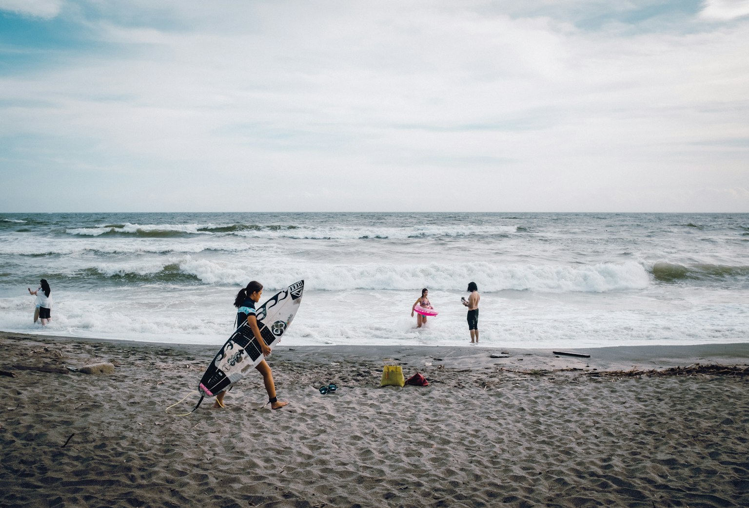 Pemandangan pantai dengan seseorang membawa papan selancar dan lainnya bermain di ombak