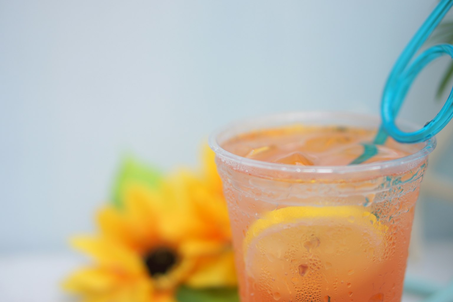 A cup of cold drink with lemon slices and a blue straw alongside a sunflower
