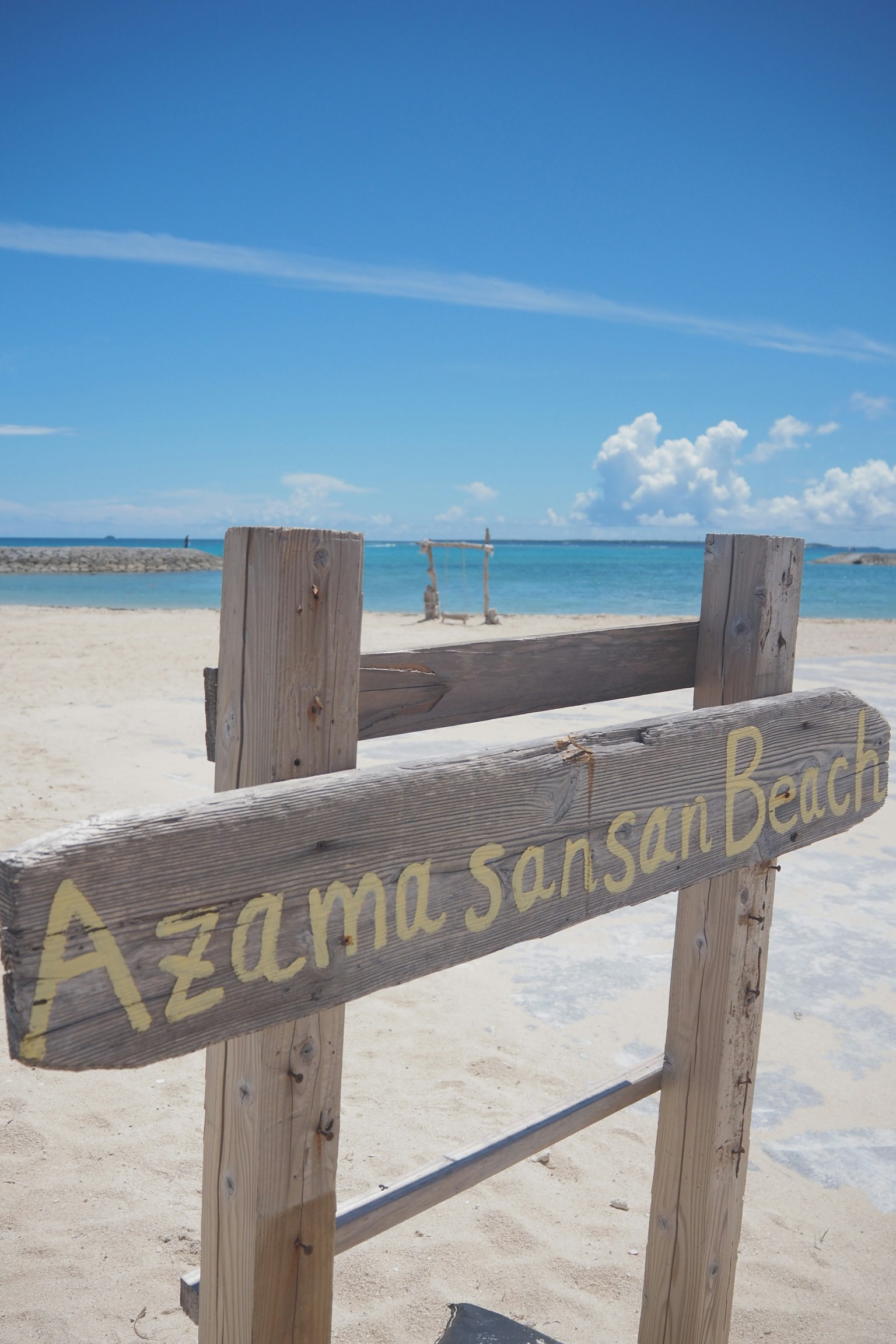 ป้ายไม้สำหรับ Azama sansan Beach กับพื้นหลังท้องฟ้าสีฟ้าและมหาสมุทร