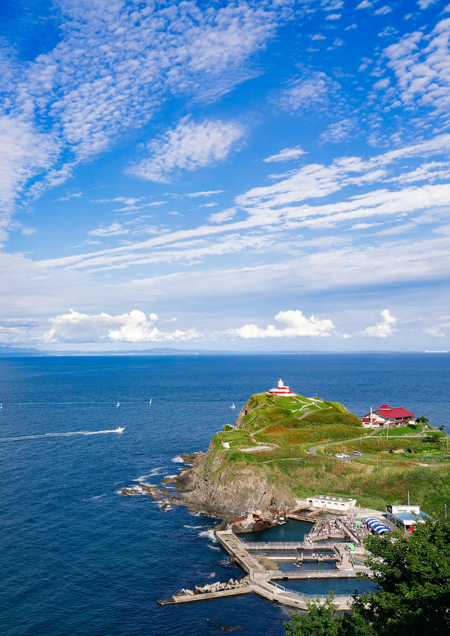 Colina verde con un faro y edificios rodeados de cielo azul y océano