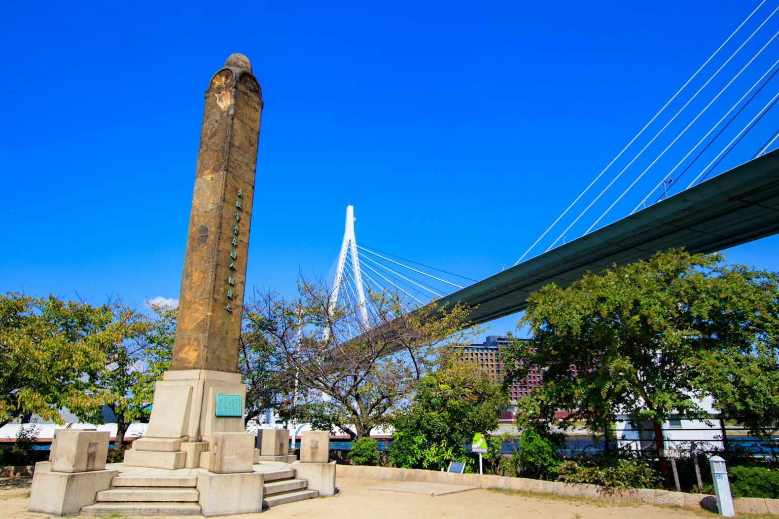 Monumen batu kuno di bawah langit biru cerah dengan jembatan gantung modern di latar belakang
