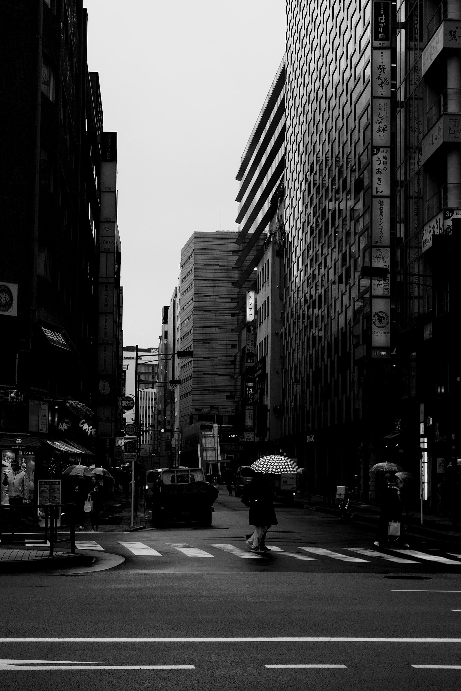 Paisaje urbano en blanco y negro con una persona que lleva un paraguas cruzando la intersección