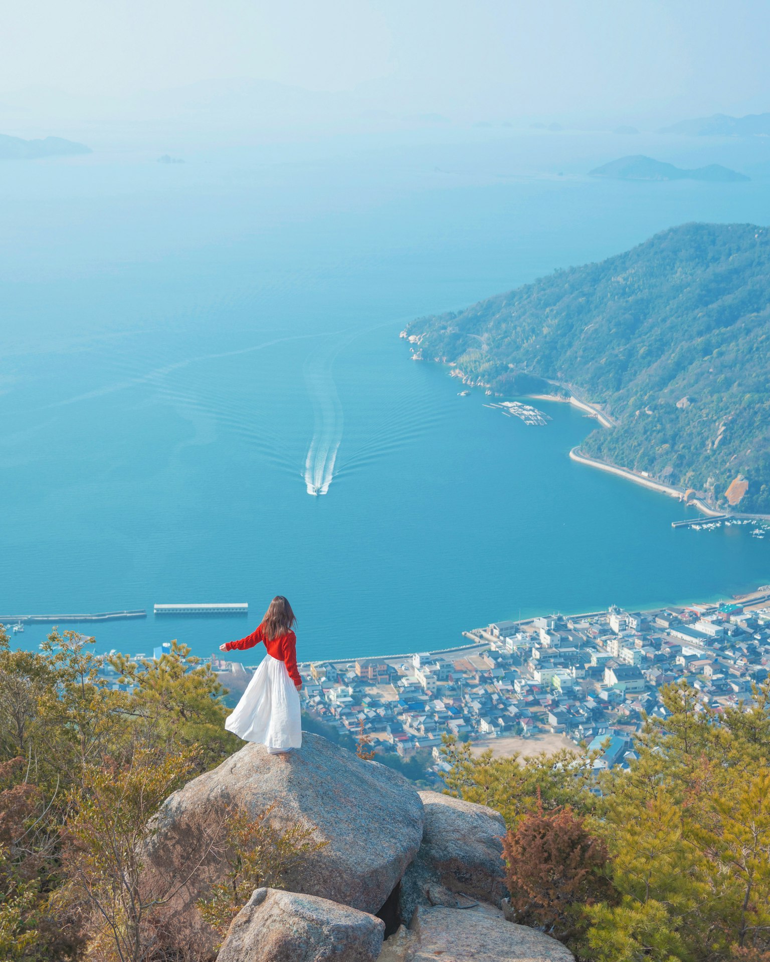 Una mujer con blusa roja y falda blanca de pie sobre una roca con vista al mar
