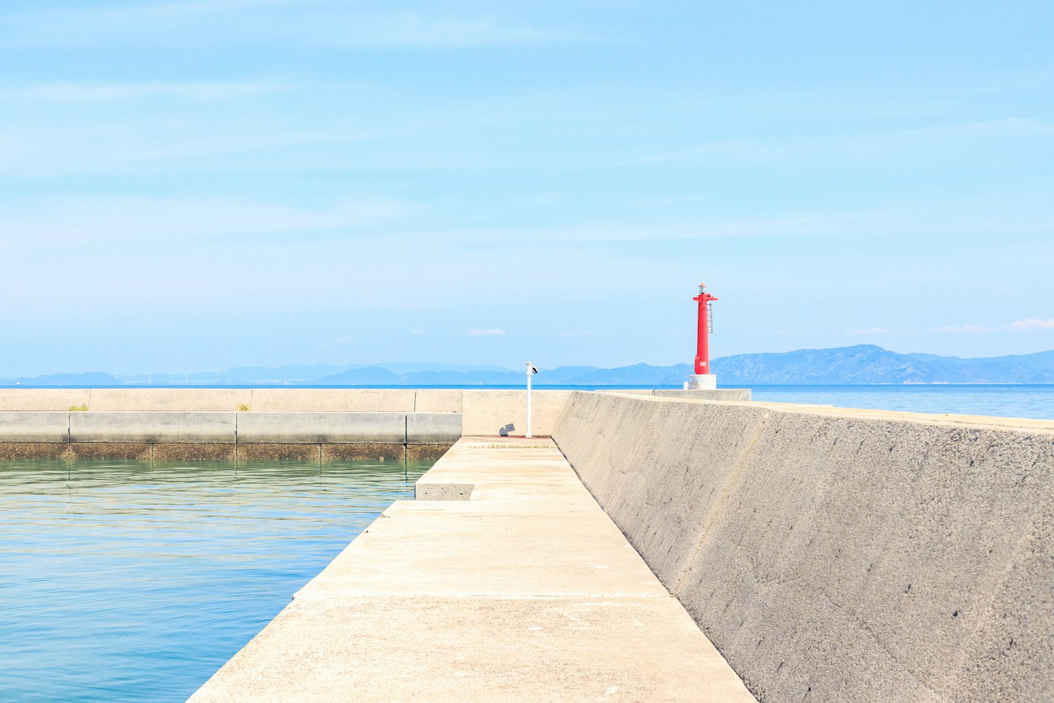 Muelle de concreto rodeado de cielo azul y agua con un faro rojo