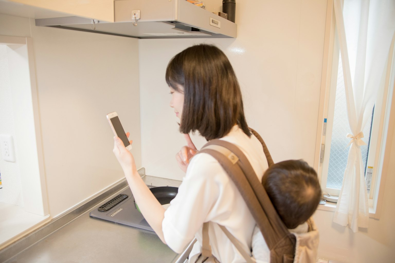 Mère utilisant un smartphone dans la cuisine avec un enfant sur le dos