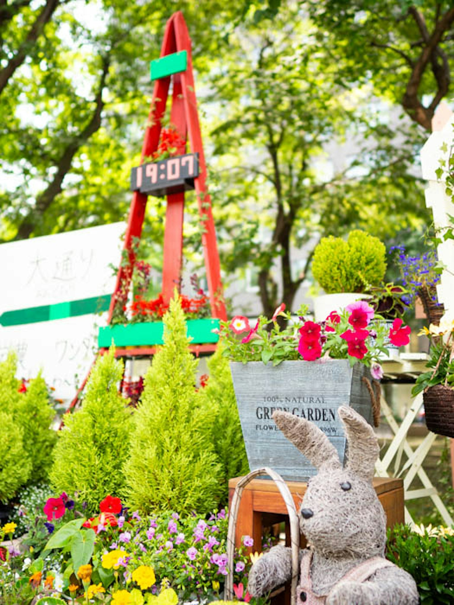 Bunte Blumen und grüne Sträucher umgeben eine Gartenszene mit einer roten Uhrenstruktur und einem Stoffhasen