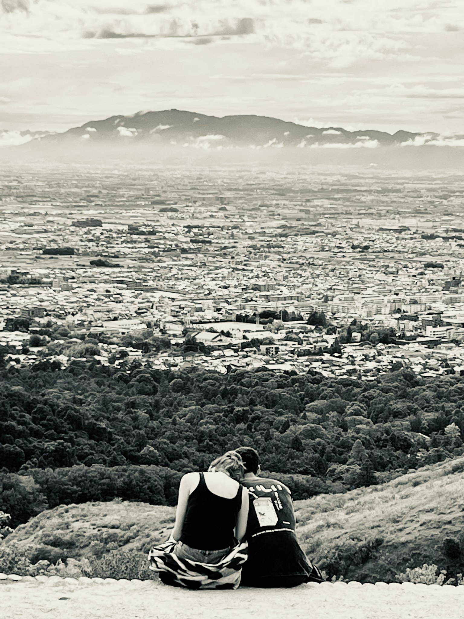 二人の人物が山の頂上で座っている風景のモノクロ写真 彼らの後ろには広がる都市の景色が見える