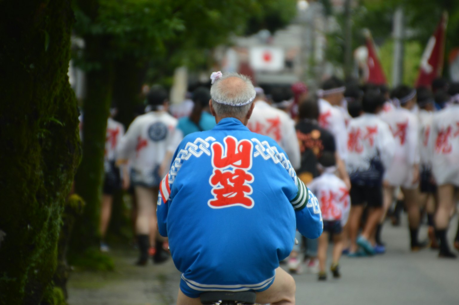 Un uomo anziano in giacca blu è seduto di spalle La sua giacca presenta caratteri kanji e persone che partecipano a un festival sono visibili sullo sfondo