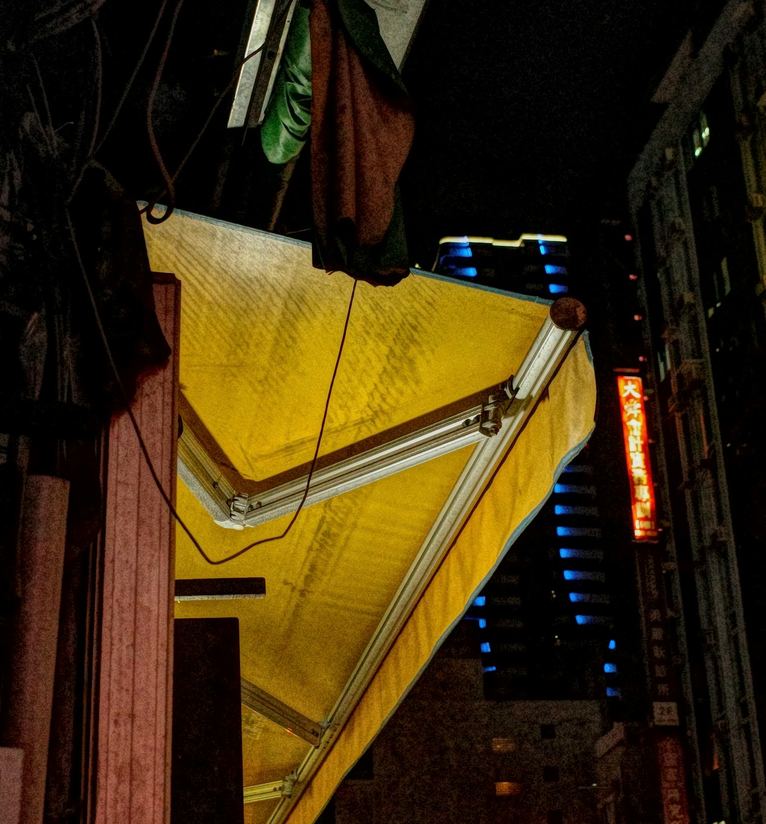 夜の都市の風景で、黄色いテントが目立つ。背景には青い光を放つビルが見える