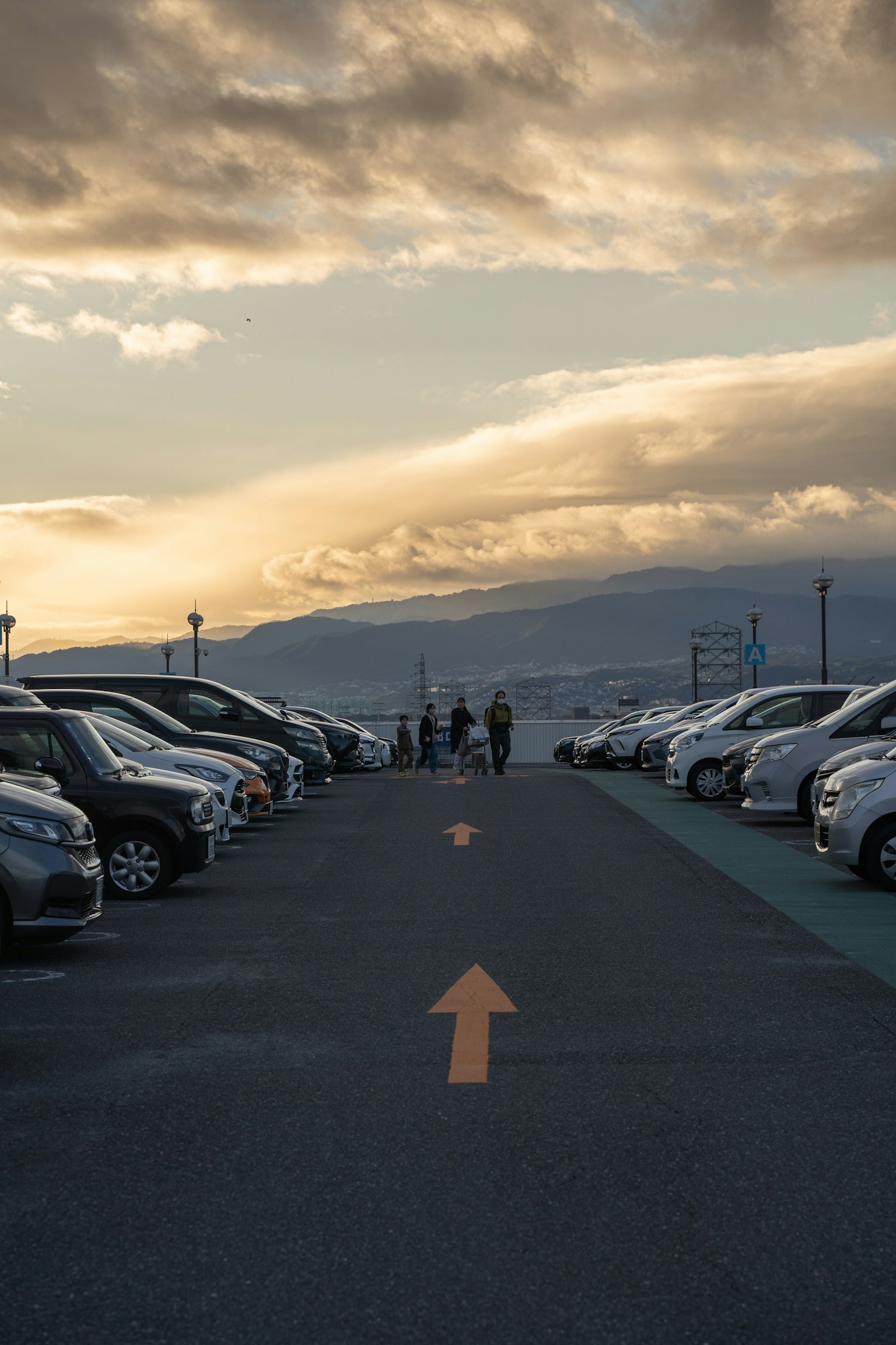 File di auto in un parcheggio con persone che camminano sotto un cielo al tramonto