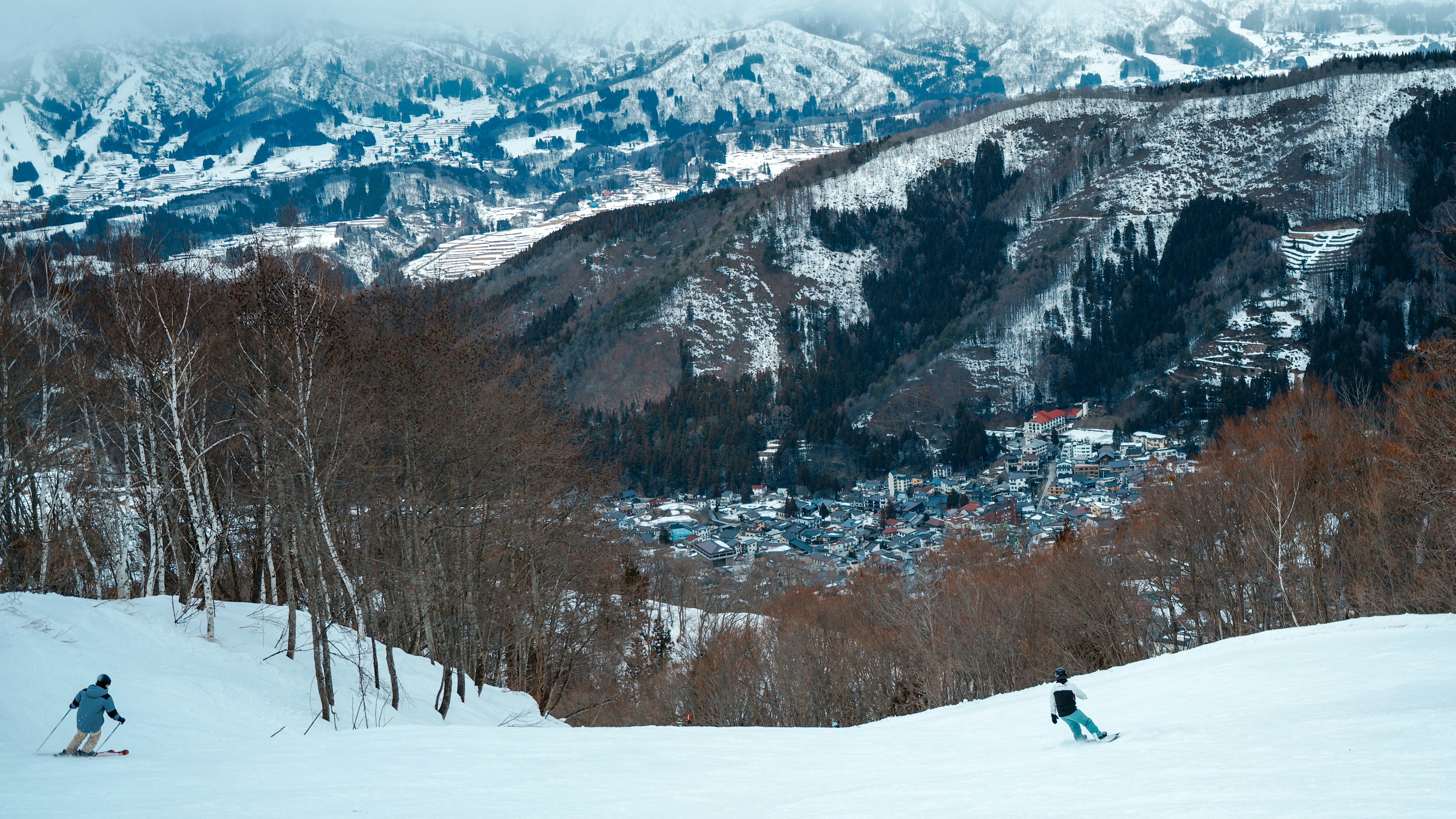 雪に覆われた山の斜面でスキーをする二人のスキーヤーと遠くの町の景色