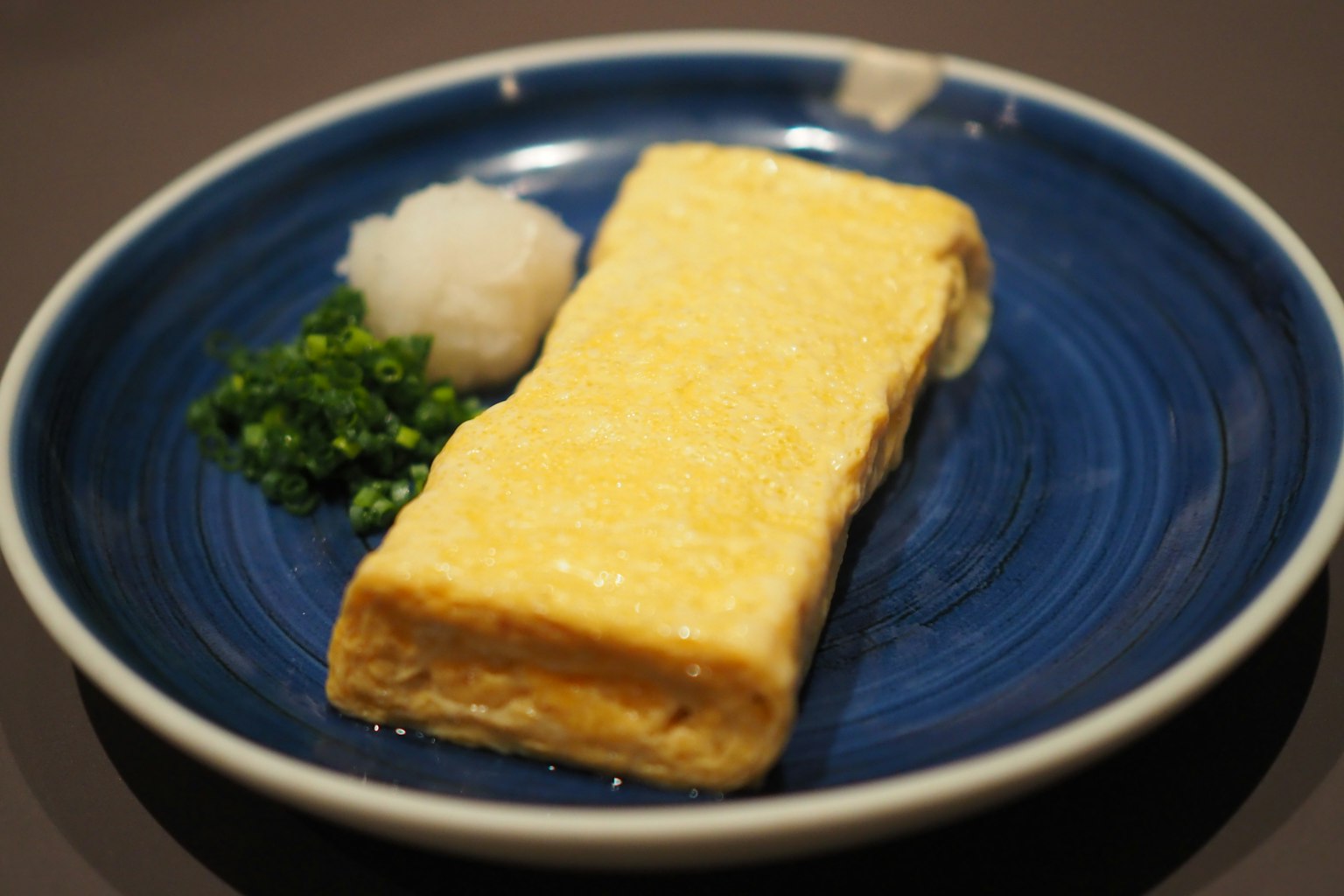 Omelet Jepang tebal disajikan di piring biru dengan lobak parut dan daun bawang cincang
