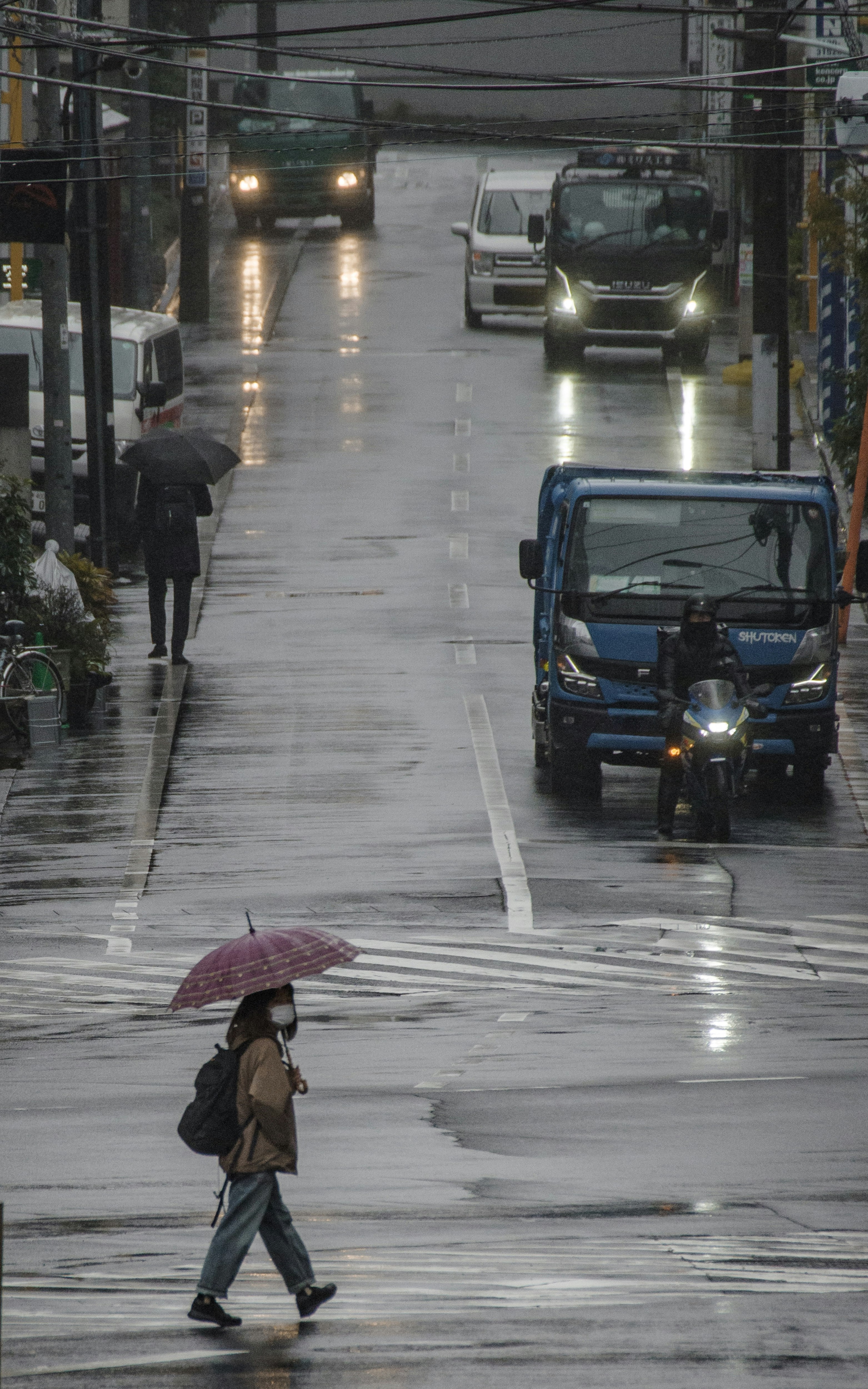 雨の東京の街並みを傘をさして歩く人と車のある路地