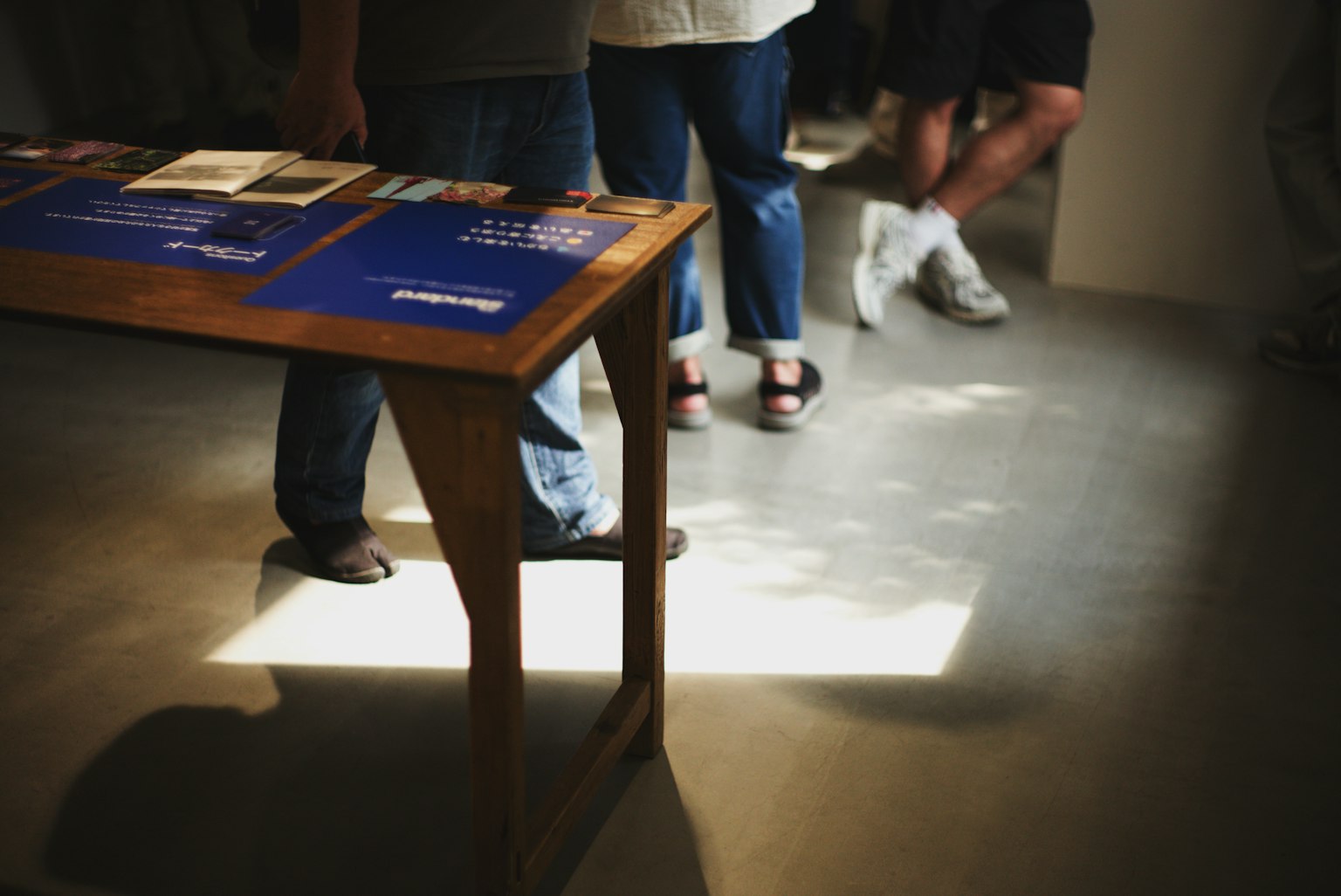 Pies de personas en un espacio de exhibición con una mesa de madera que muestra materiales azules
