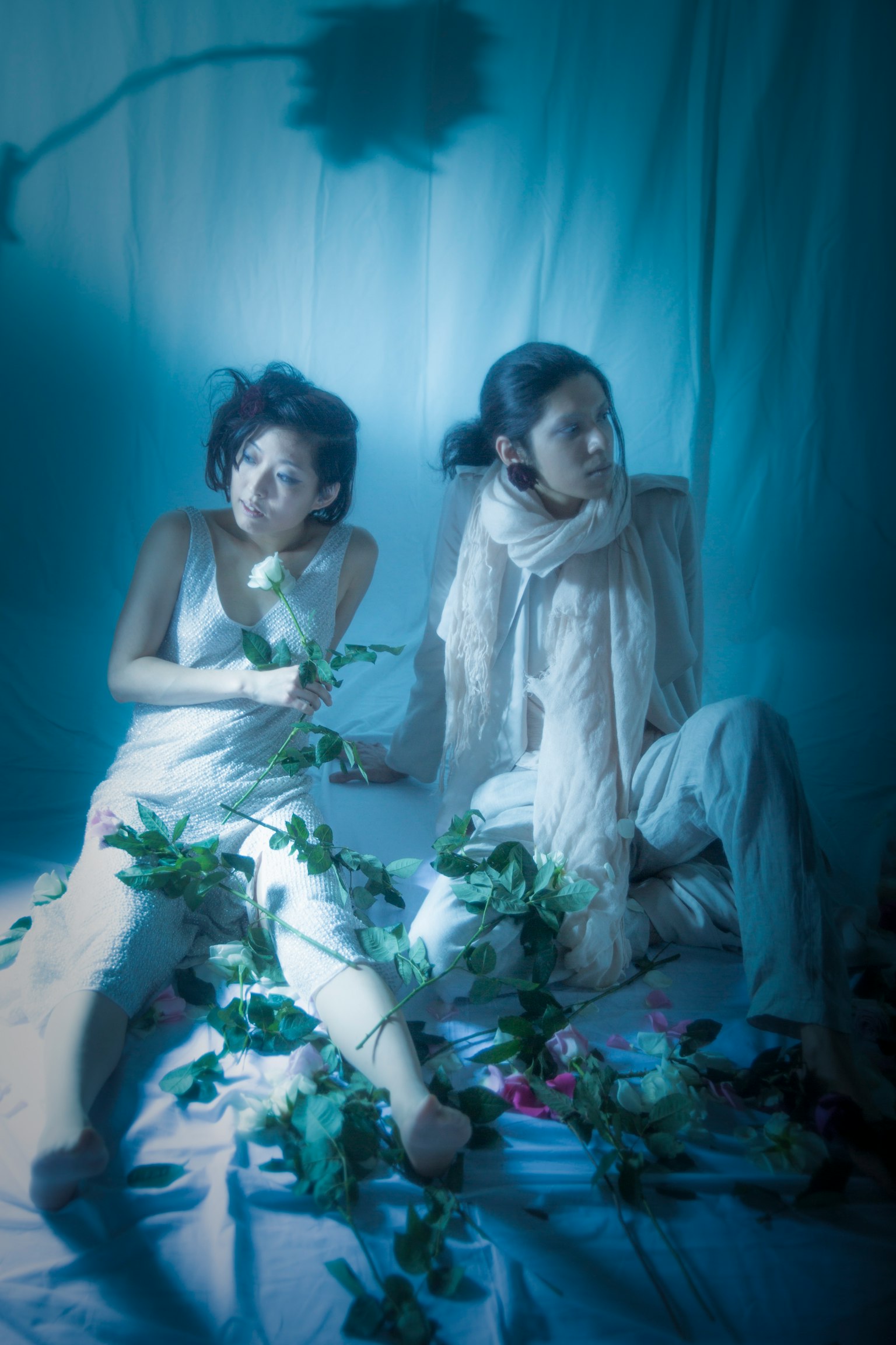 两位穿着白色衣服的女性坐在蓝色背景前 一位手持玫瑰 另一位围着围巾 周围散落着叶子和花朵