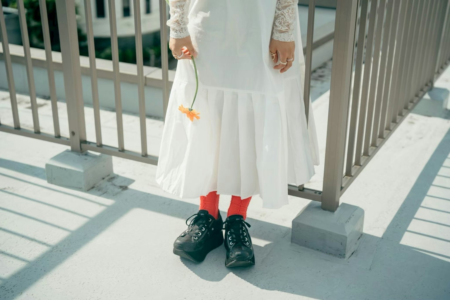 白いドレスを着た人物が赤い靴下を履いている写真で、手に花を持ち、フェンスの前に立っている