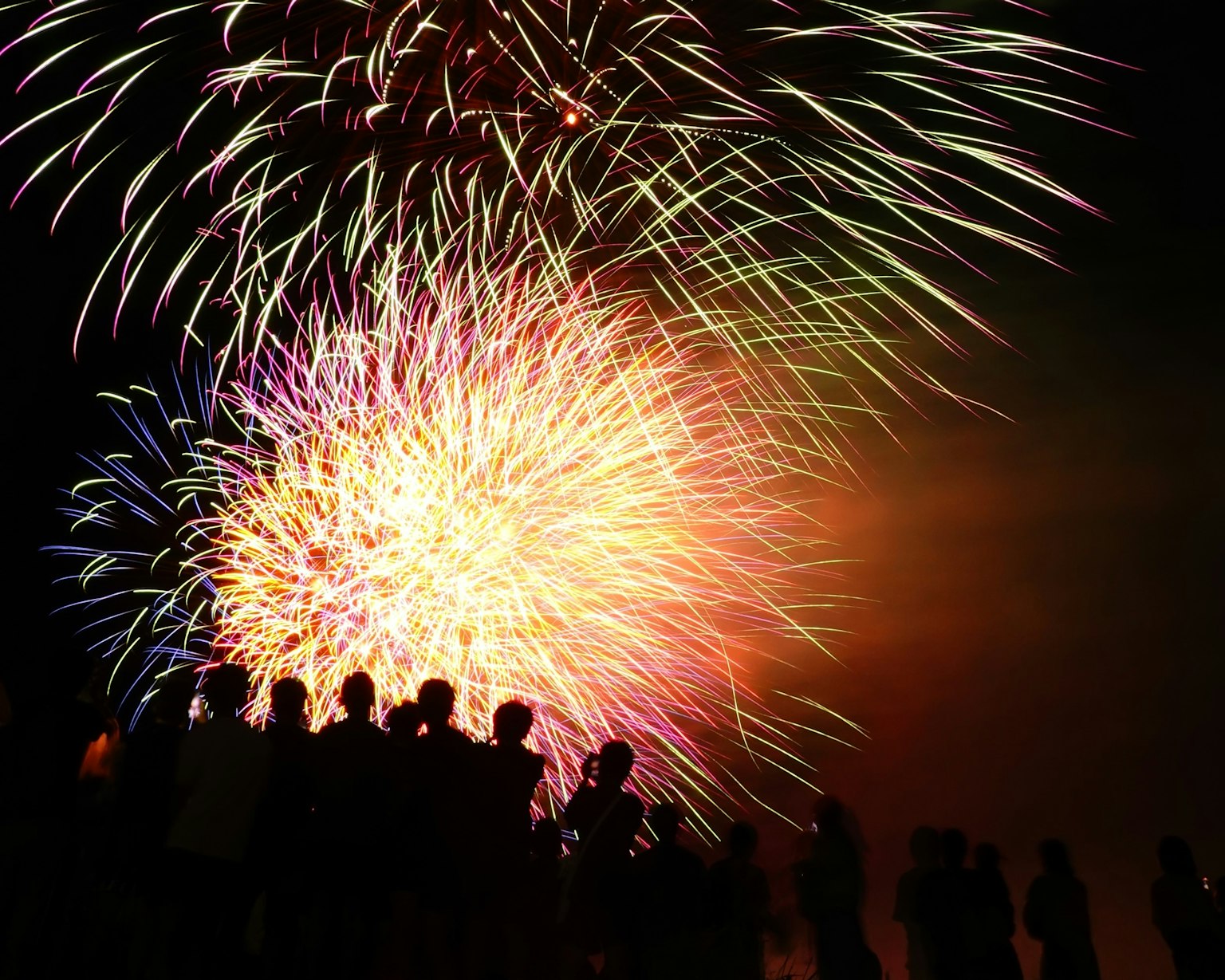 Fuegos artificiales estallando en colores vibrantes contra un cielo nocturno con siluetas de personas