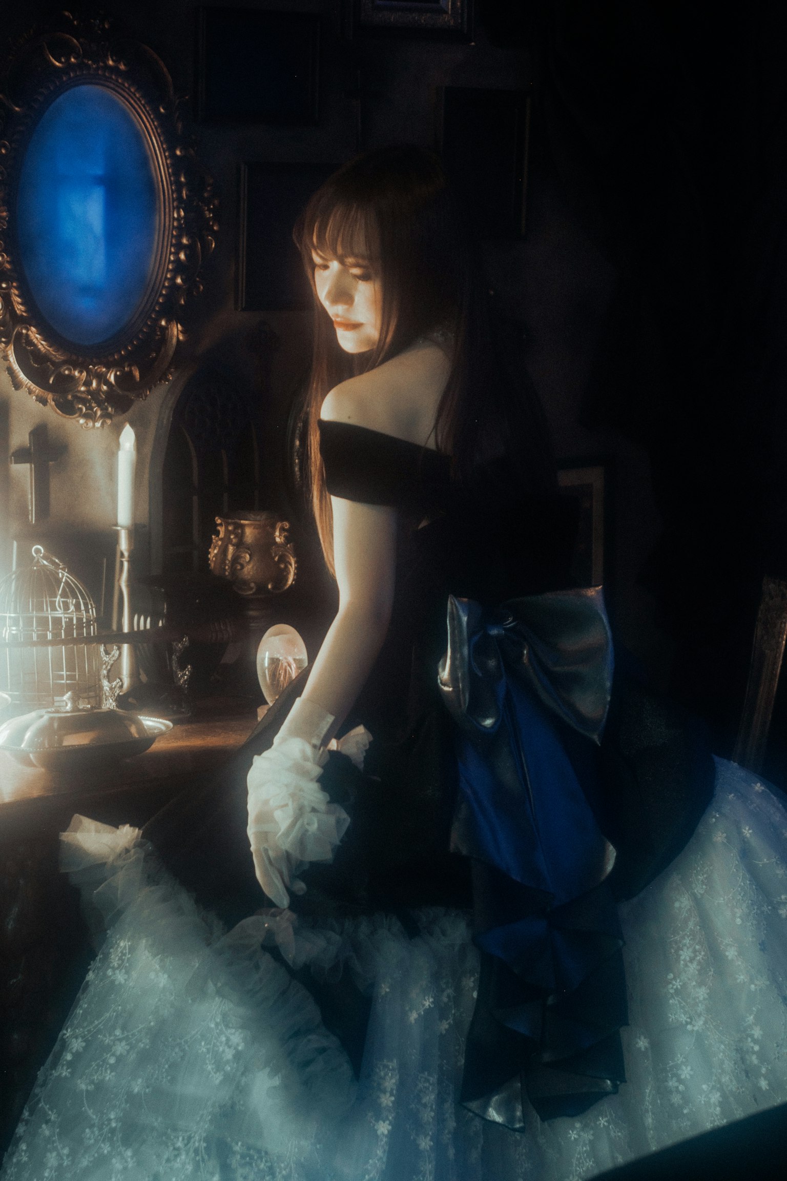 Portrait of a woman standing in front of a mirror in a dim room wearing a black top and white skirt with a blue background mirror and candlelight