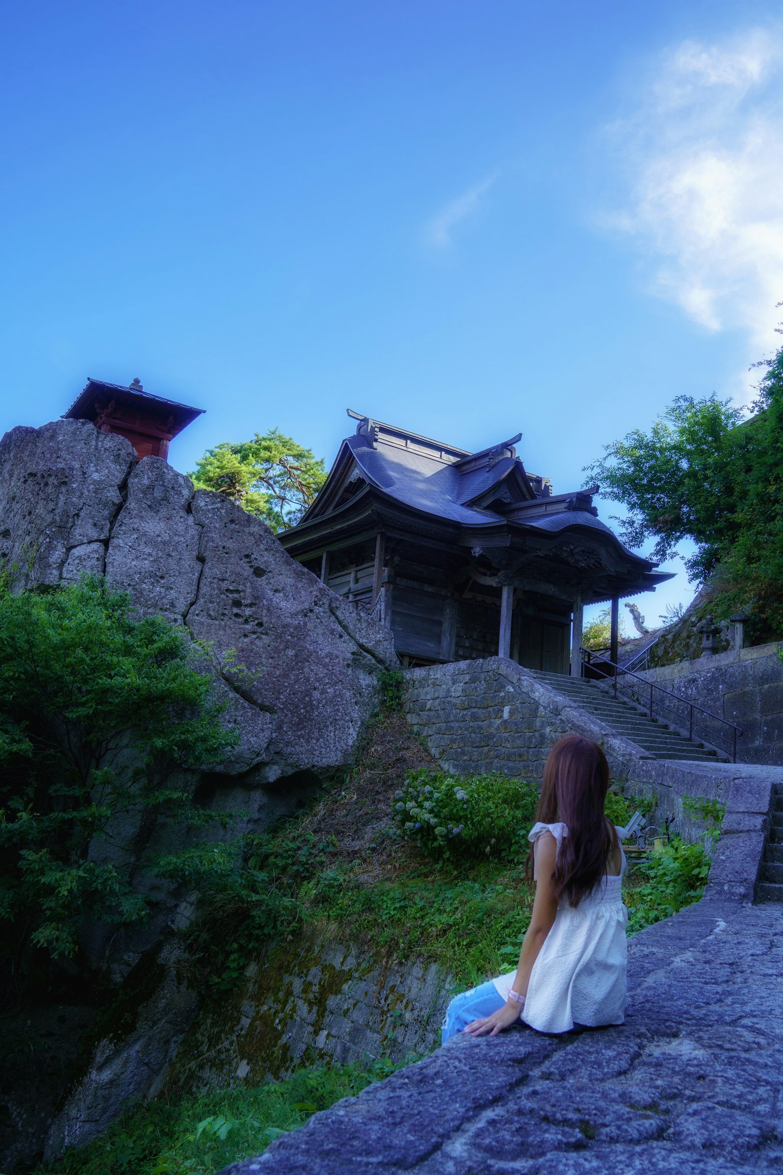 女性が岩の上に座り、神社を見上げている風景 青空と緑の木々が背景