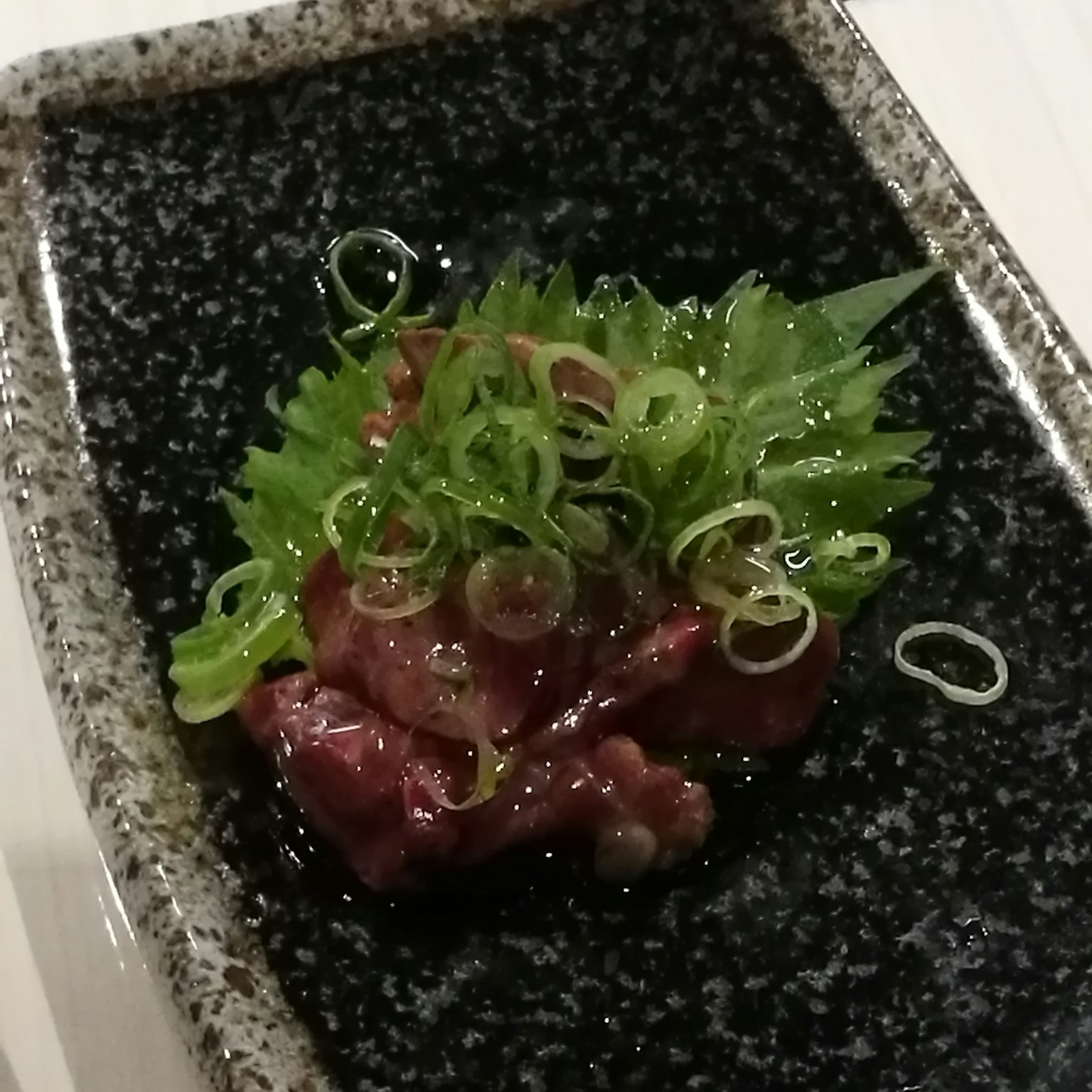 Hidangan daging mentah segar yang dihias dengan bawang hijau cincang halus dan sayuran daun di atas piring hitam