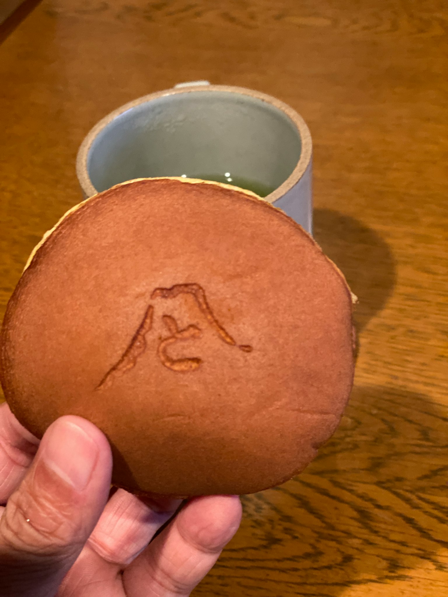 Une main tenant un wagashi en forme de montagne Fuji avec du matcha en arrière-plan