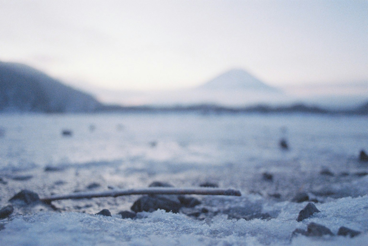 Close-up of pebbles and a twig on a snow-covered lakeshore with Mount Fuji in the background in winter scenery