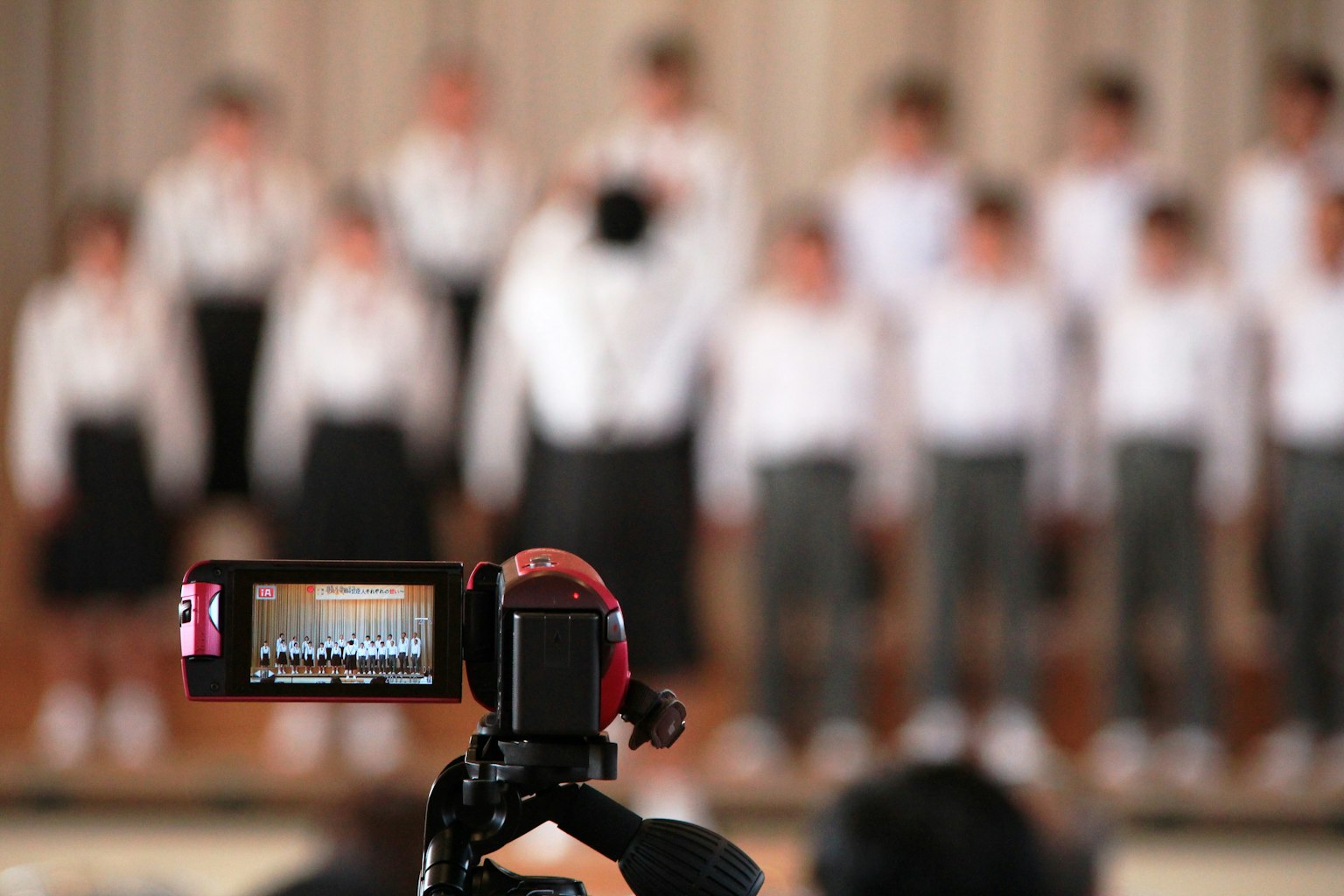 Una cámara filmando una actuación de coro con estudiantes en camisas blancas y faldas al fondo