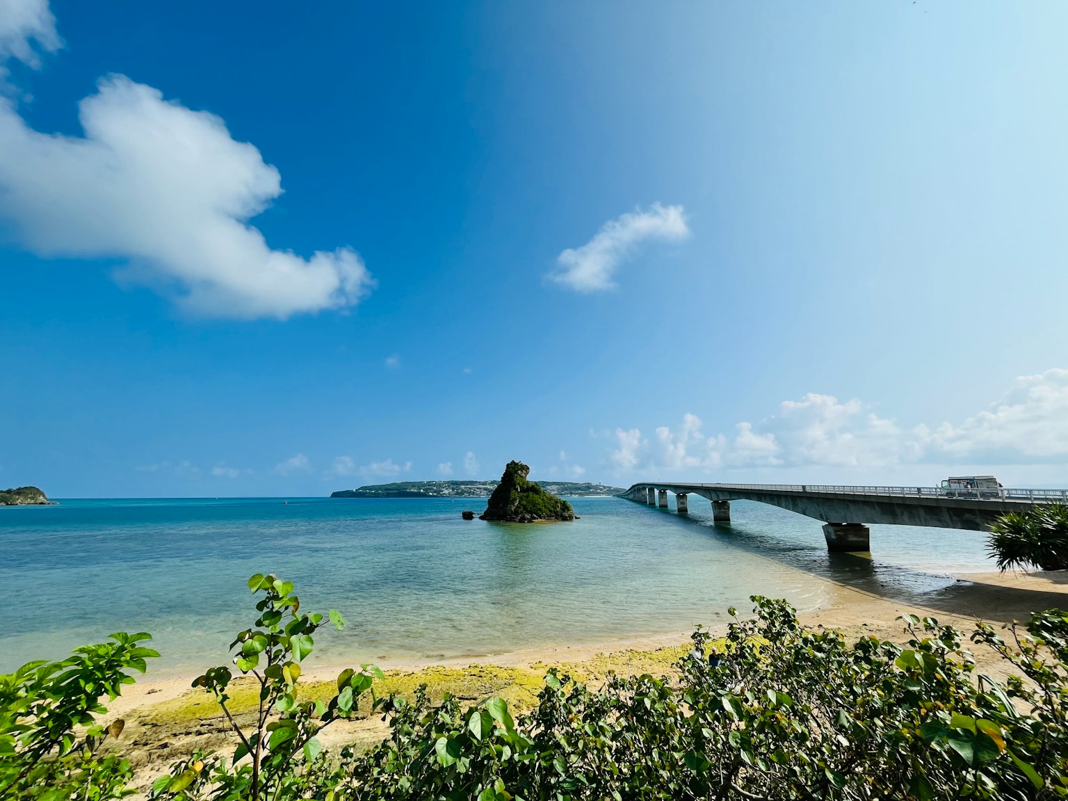 Vue pittoresque d'une plage avec un pont et une île rocheuse sous un ciel bleu clair