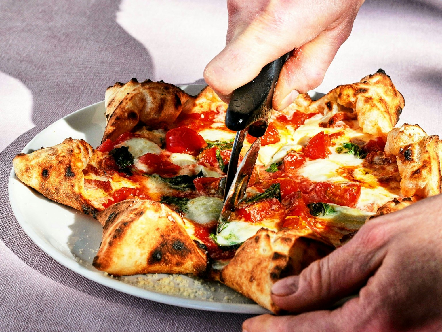 Eine Hand schneidet eine Pizza mit bunten Belägen auf einem weißen Teller