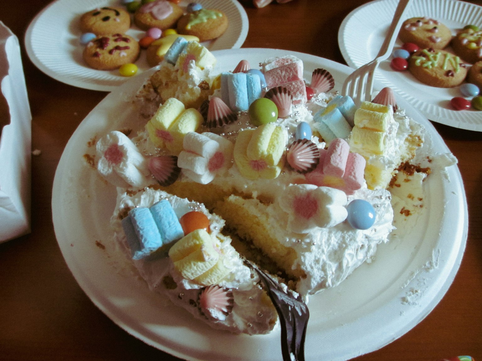 A slice of cake with colorful toppings and plates of cookies in the background