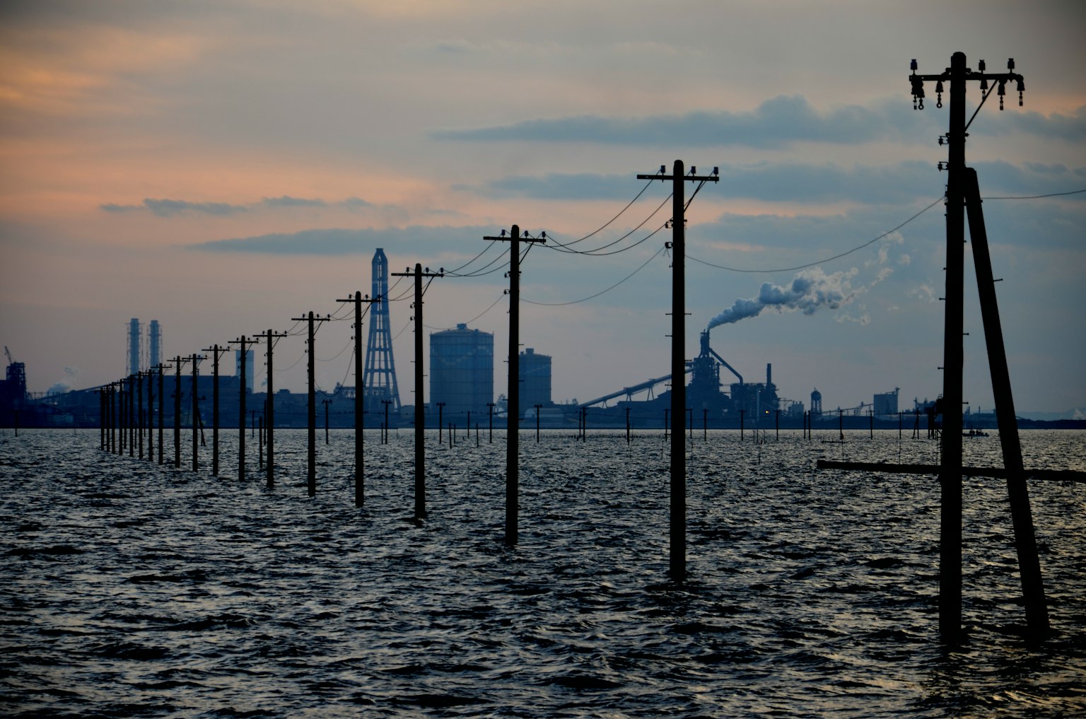 Silhouetted power poles in water with factory skyline at sunset