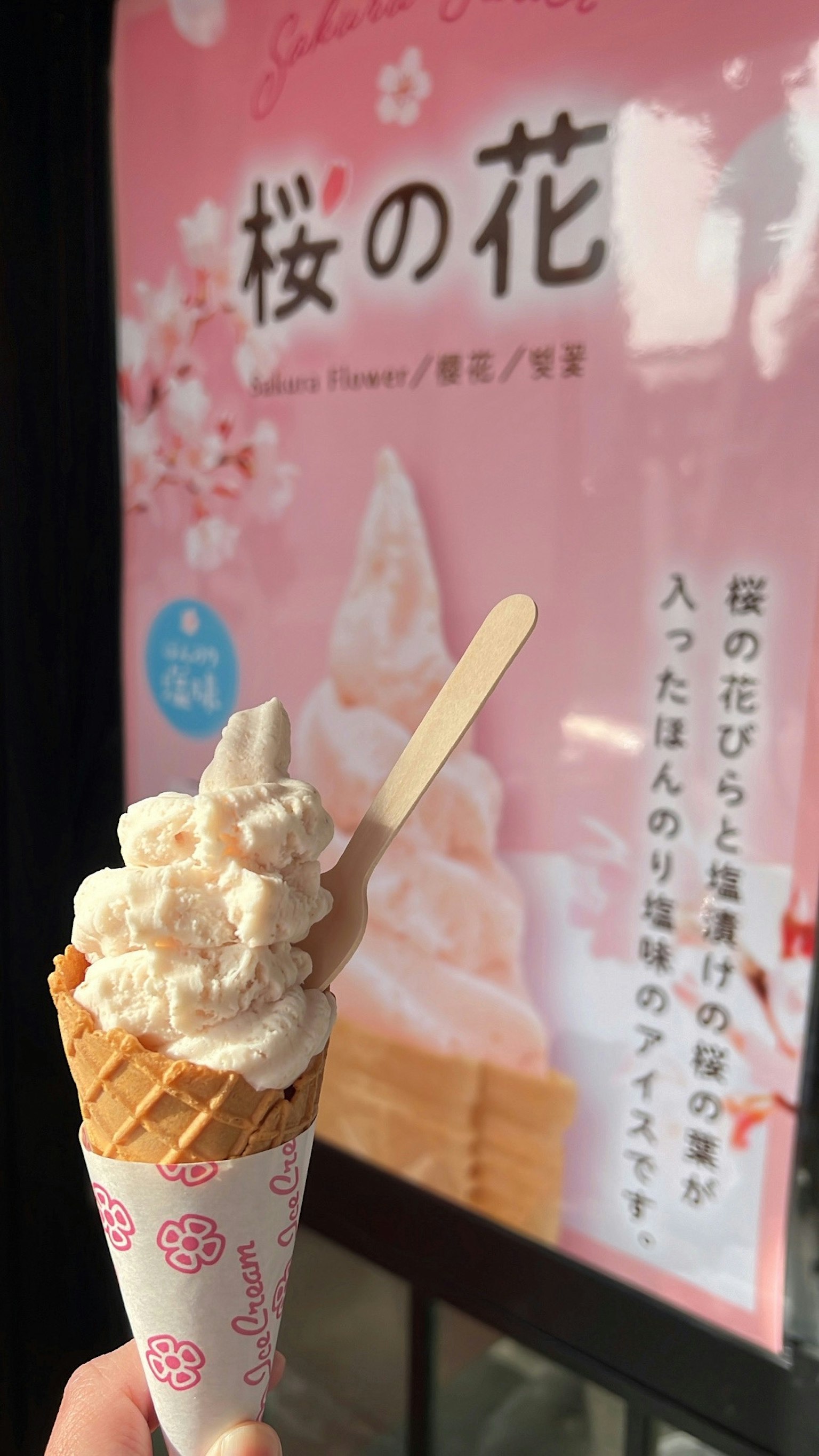 Tangan memegang kerucut es krim bunga sakura dengan poster bunga sakura di latar belakang