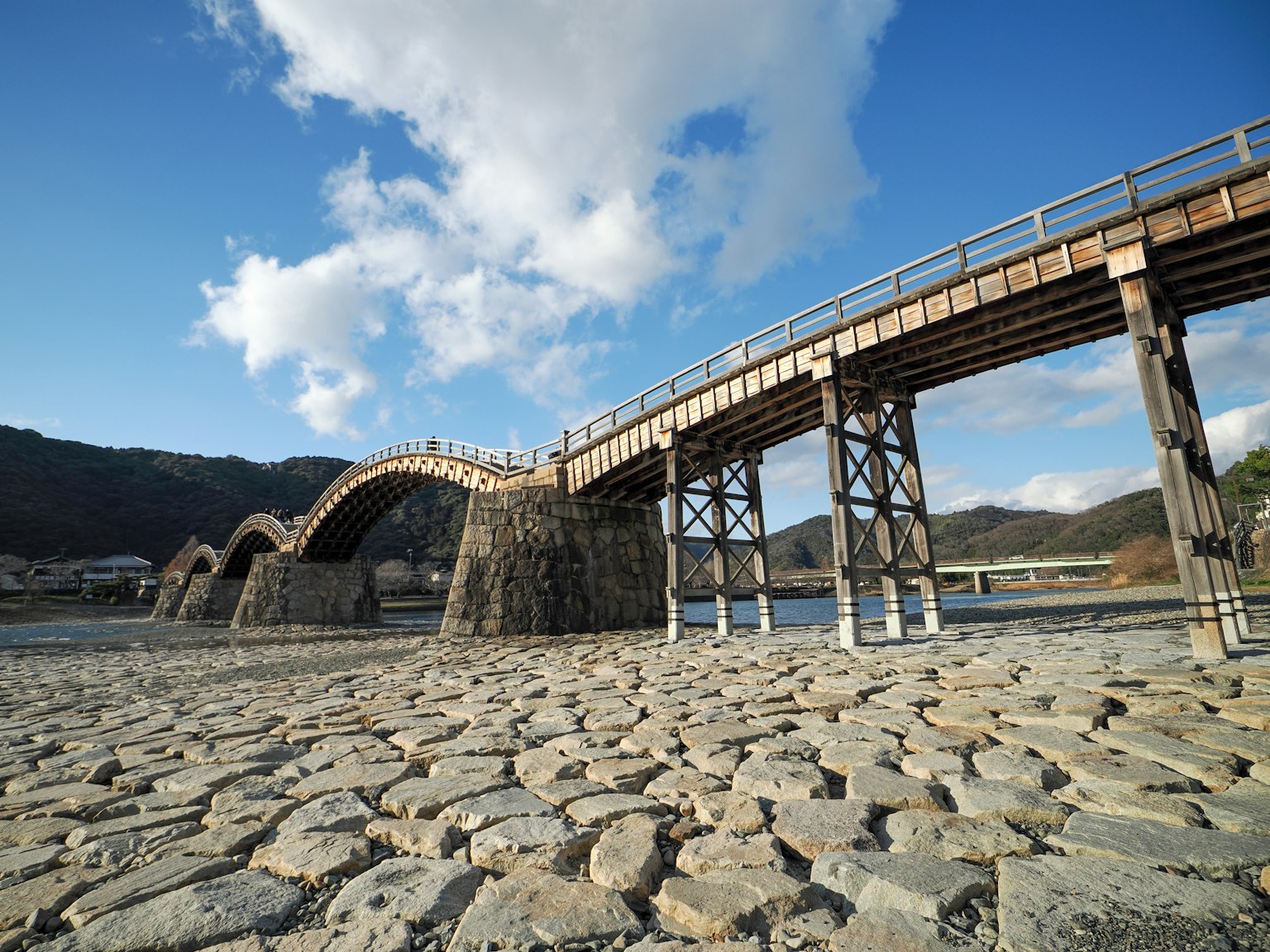 Un pont en bois en arc sous un ciel bleu avec des fondations en pierre et un lit de rivière asséché