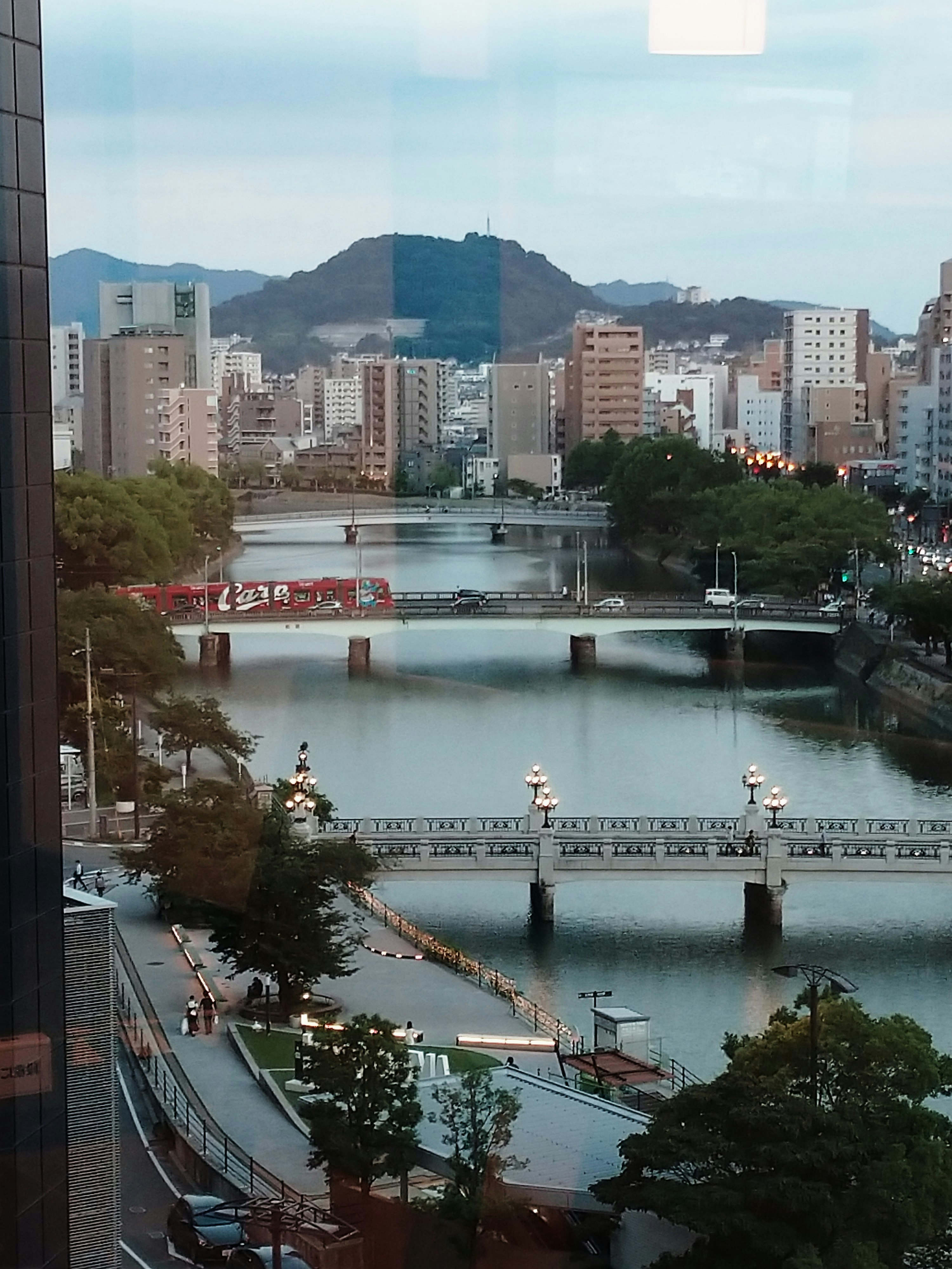 川沿いの橋と川岸の遊歩道、山並みと市街地が見える風景