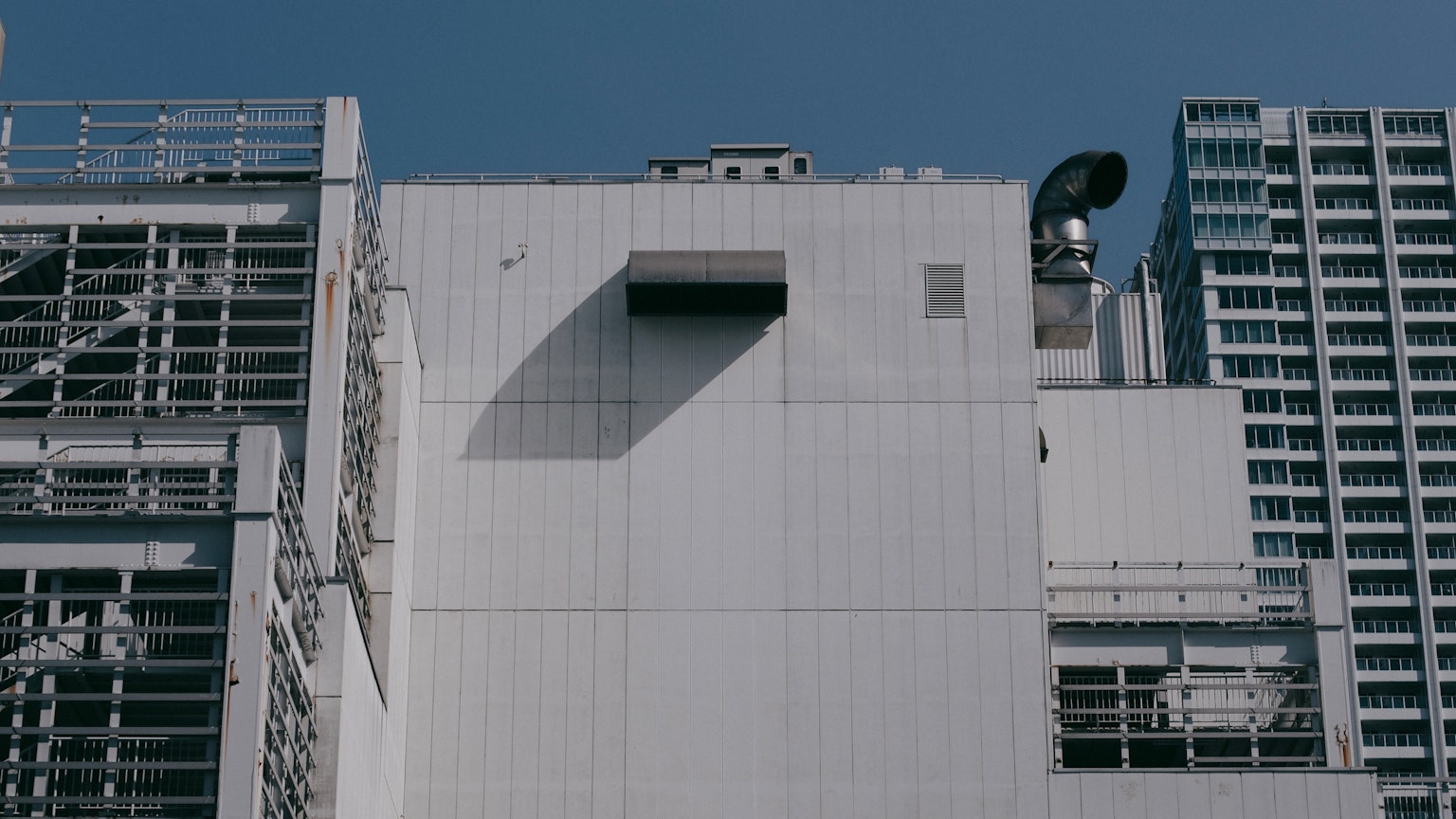 Façade d'un bâtiment moderne avec des murs blancs et des structures de tuyaux en métal sous un ciel bleu projetant des ombres