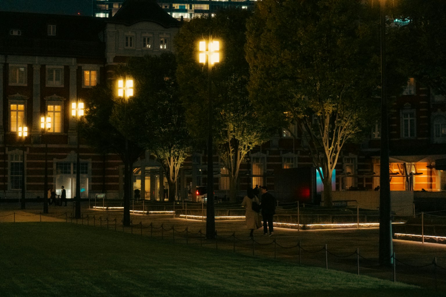 人們在夜間的公園裡散步，背景是街燈和綠色樹木