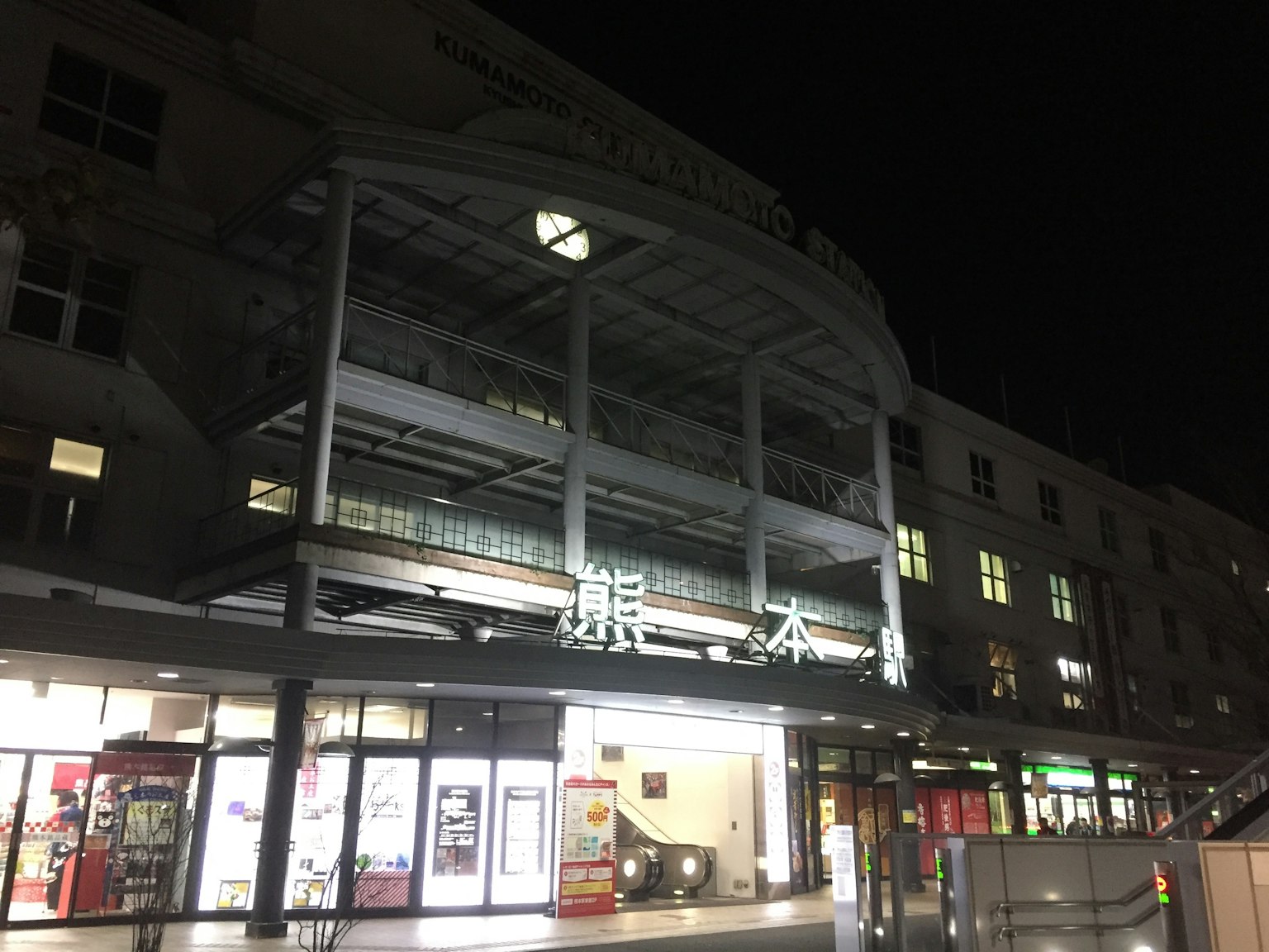 Vista exterior de la estación de Kumamoto de noche con letreros brillantes y edificios circundantes