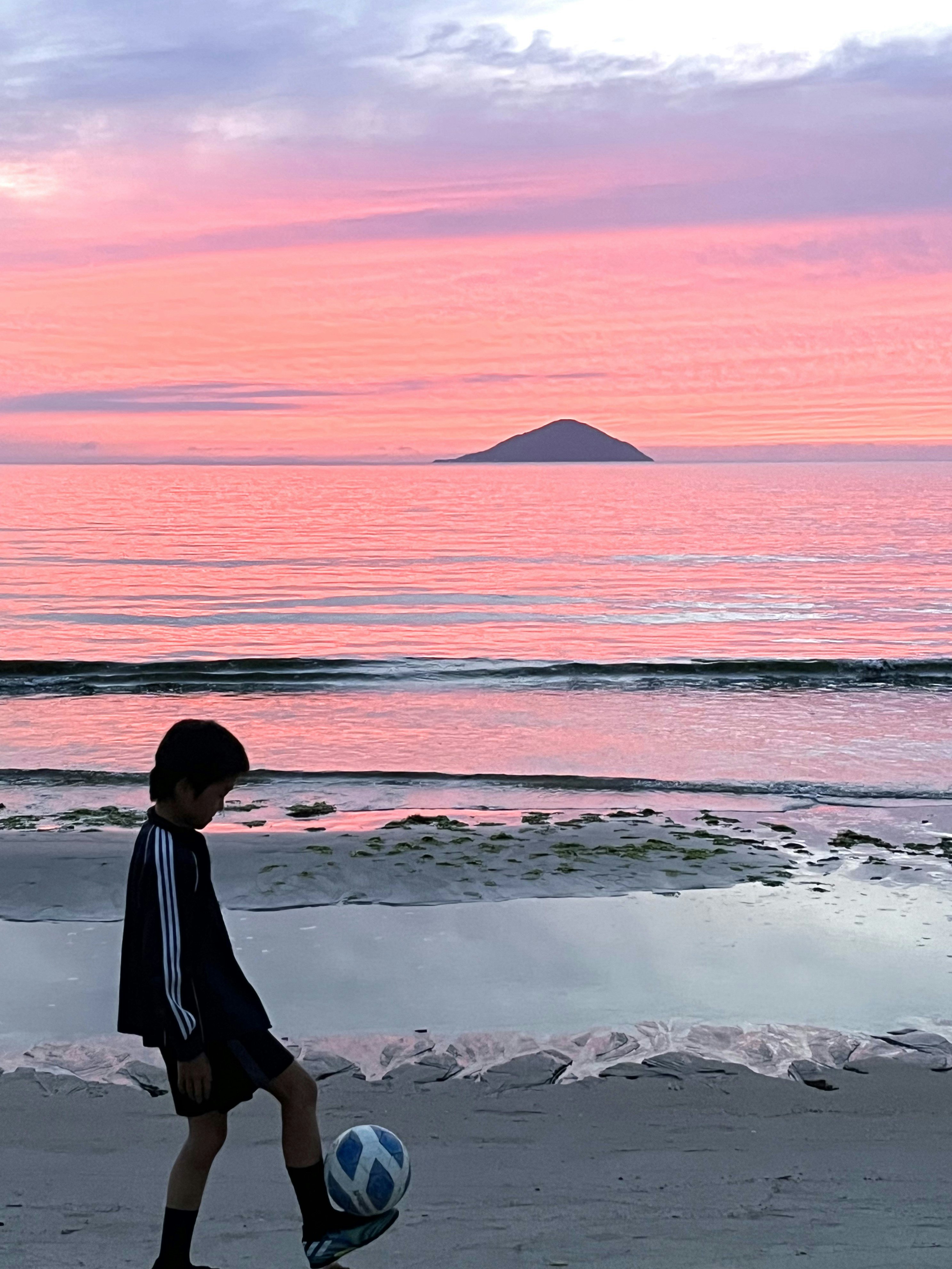 夕焼けの海辺でサッカーボールを持つ子ども