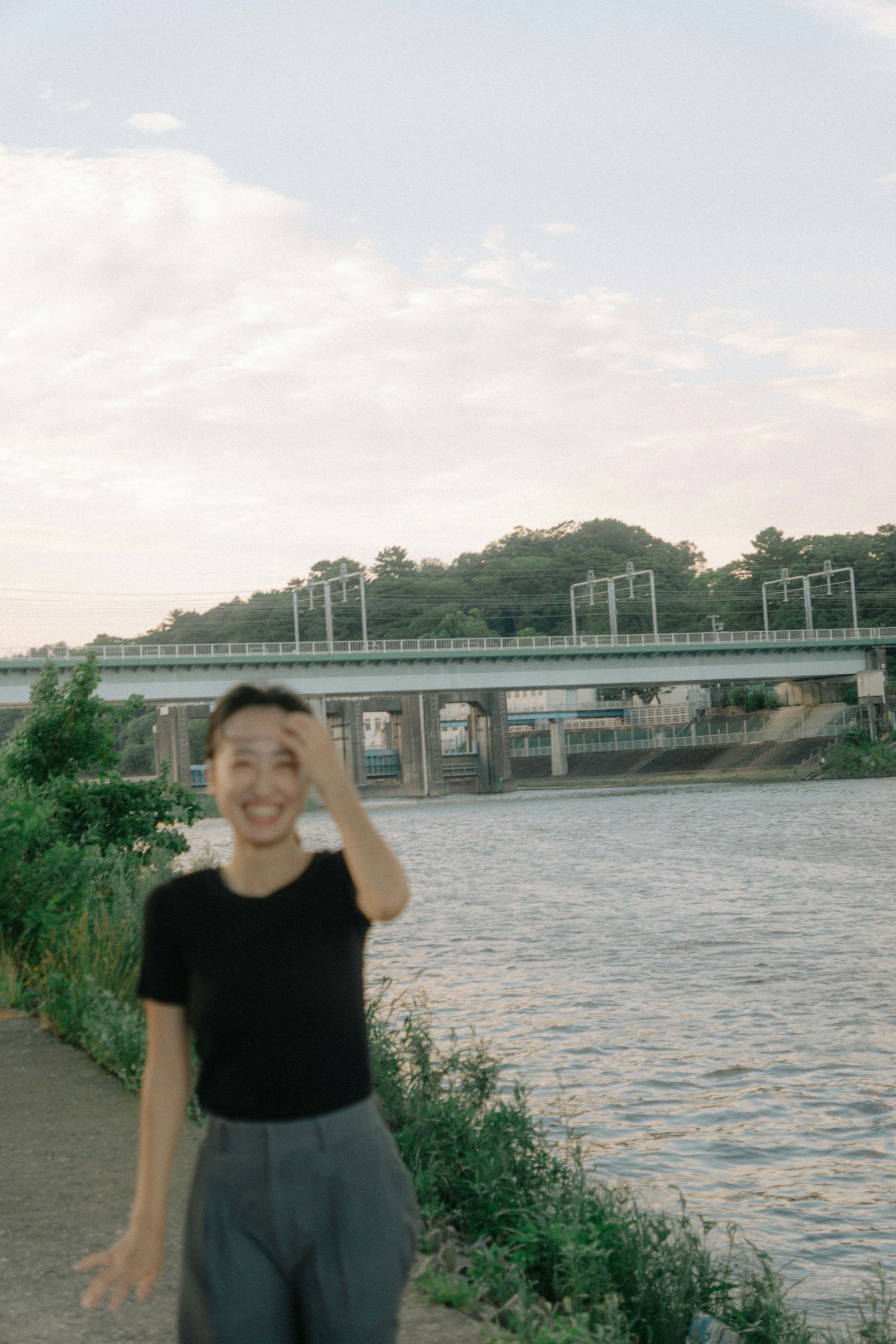 ผู้หญิงยิ้มยืนอยู่ริมแม่น้ำ มีสะพานอยู่เบื้องหลัง