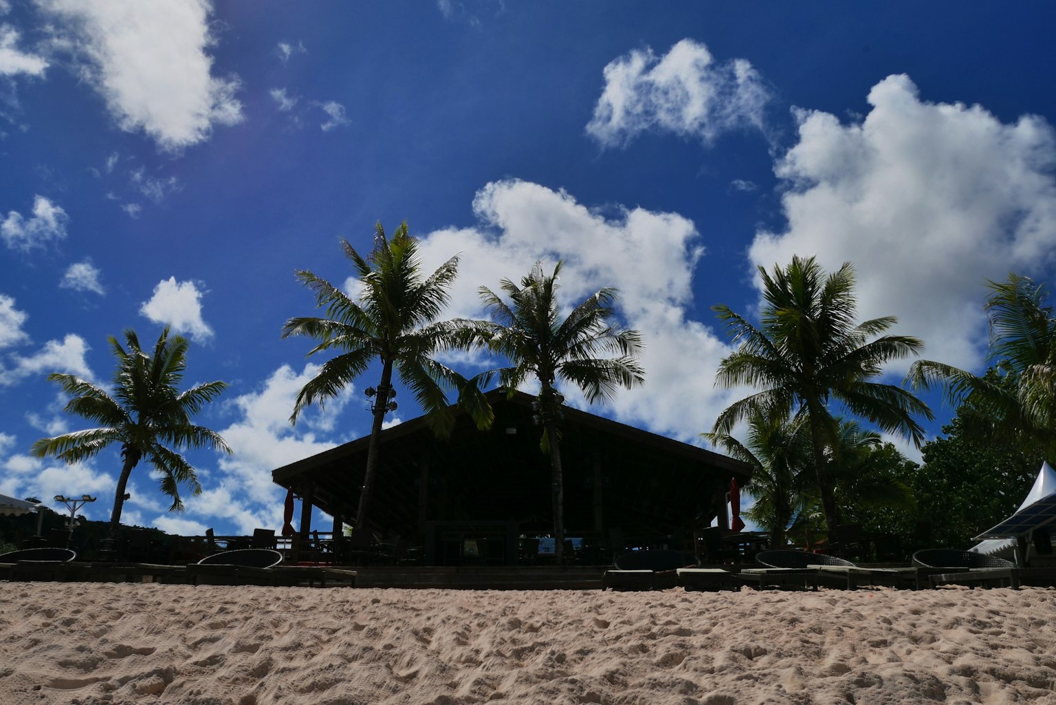 Eine Strandstruktur mit einem Reetdach umgeben von Palmen unter einem blauen Himmel und weißen Wolken