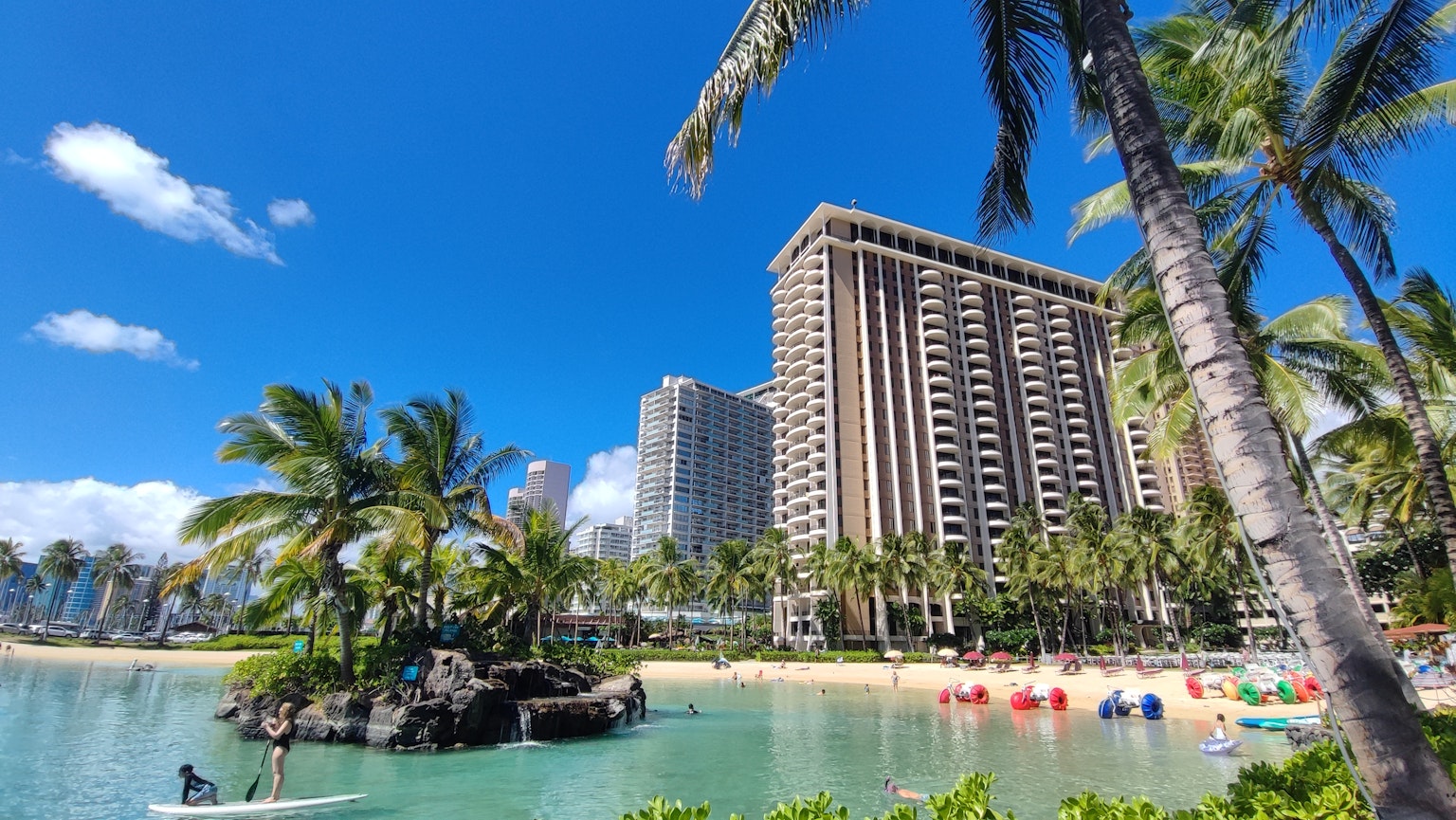 Escena de playa hawaiana con palmeras y un hotel de gran altura bajo un cielo azul claro