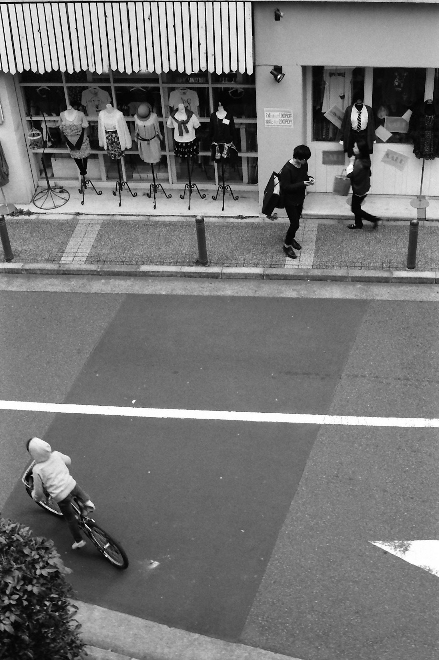 Schwarz-Weiß-Straßenszene mit einem Jungen auf einem Fahrrad und zwei Personen, die auf dem Bürgersteig gehen