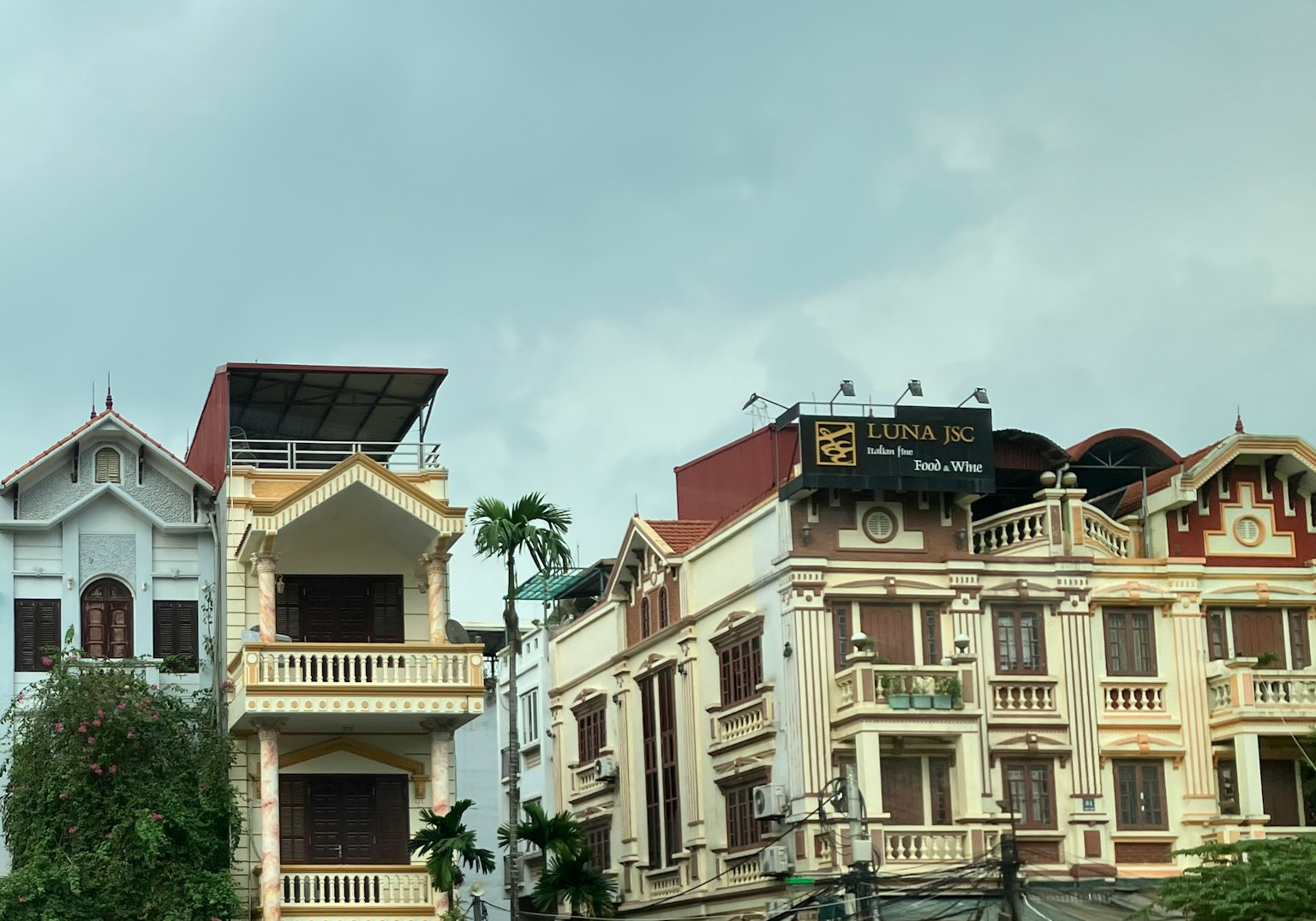 Dua bangunan berdampingan Satu berwarna putih dengan balkon berornamen Satu lagi berwarna cokelat dengan elemen dekoratif Langit mendung
