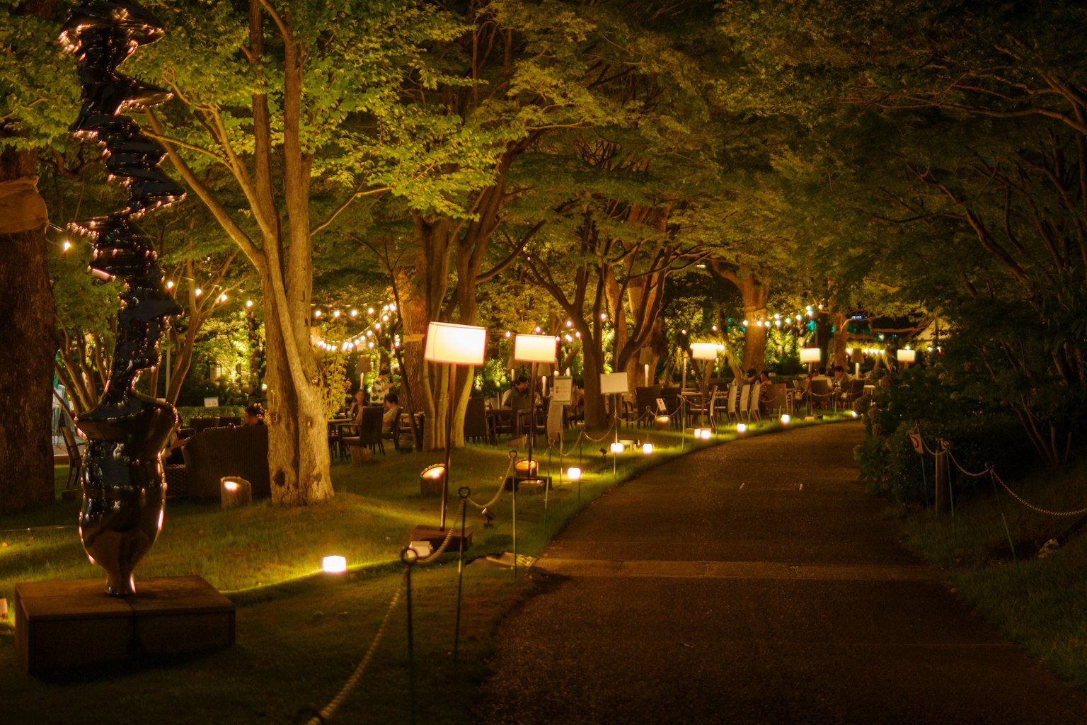 A dark pathway surrounded by green trees with bright lanterns and sculptures