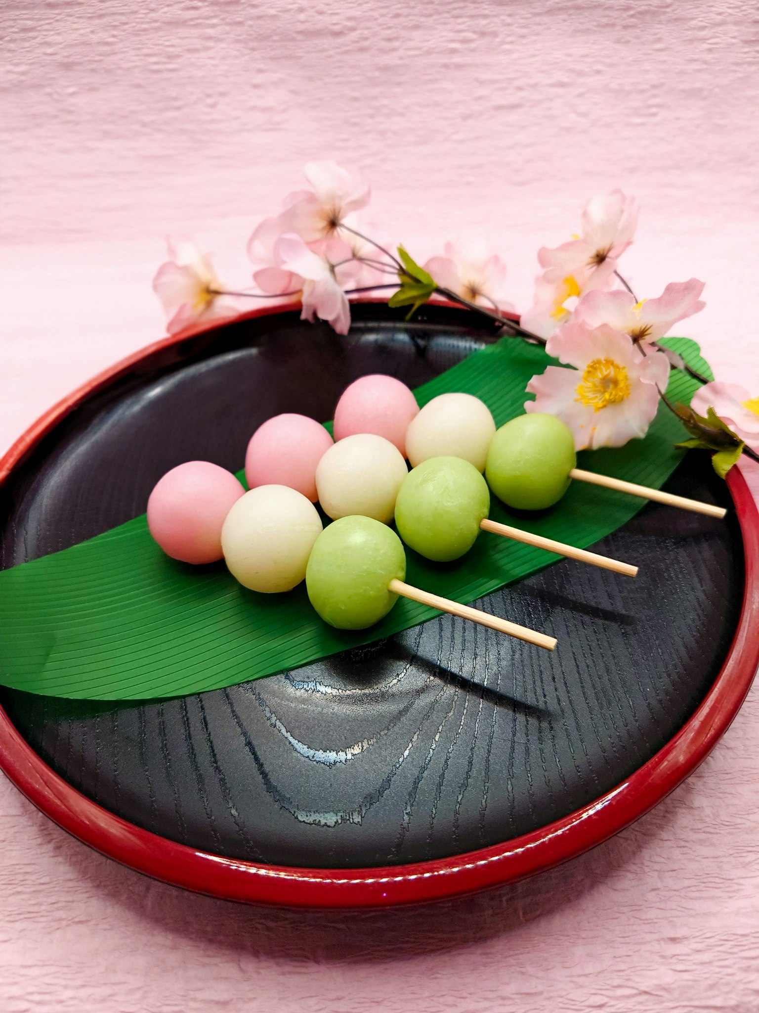 Xiên dango nhiều màu được sắp xếp trên lá xanh trên đĩa đen với hoa anh đào